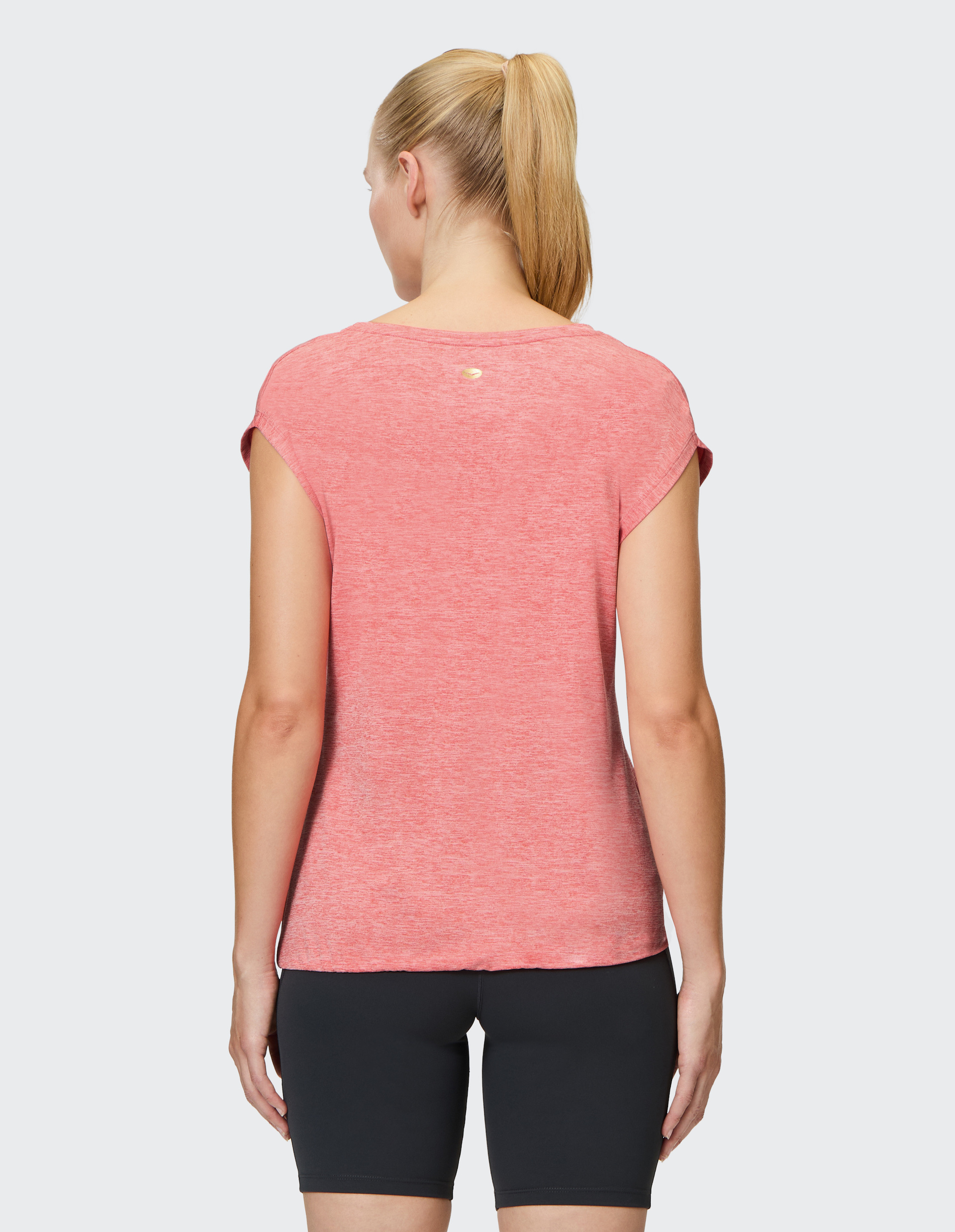 Eine Frau mit blondem Pferdeschwanz trägt ein rosafarbenes T-Shirt und schwarze Shorts, betrachtet von hinten auf grauem Hintergrund.