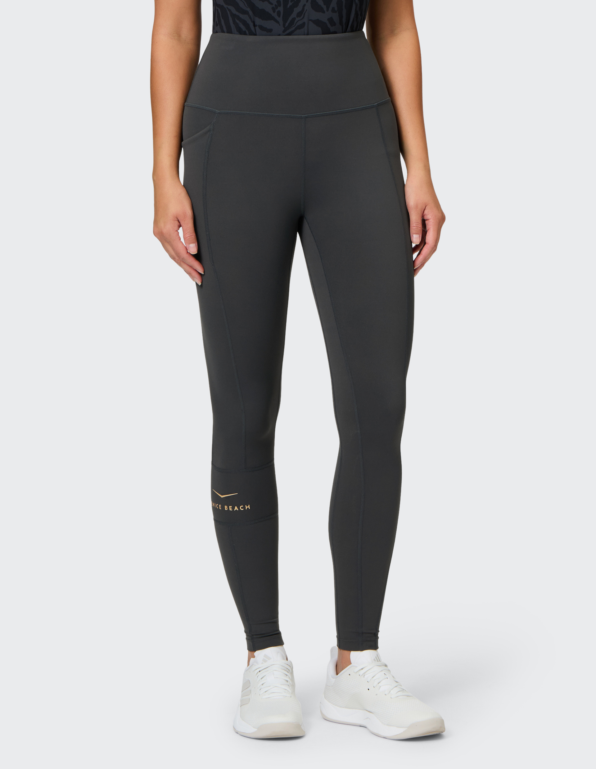 Frau in dunkler Sportleggings und weißen Sneakers, trägt ein schwarzes Oberteil mit Muster, steht auf hellem Hintergrund.