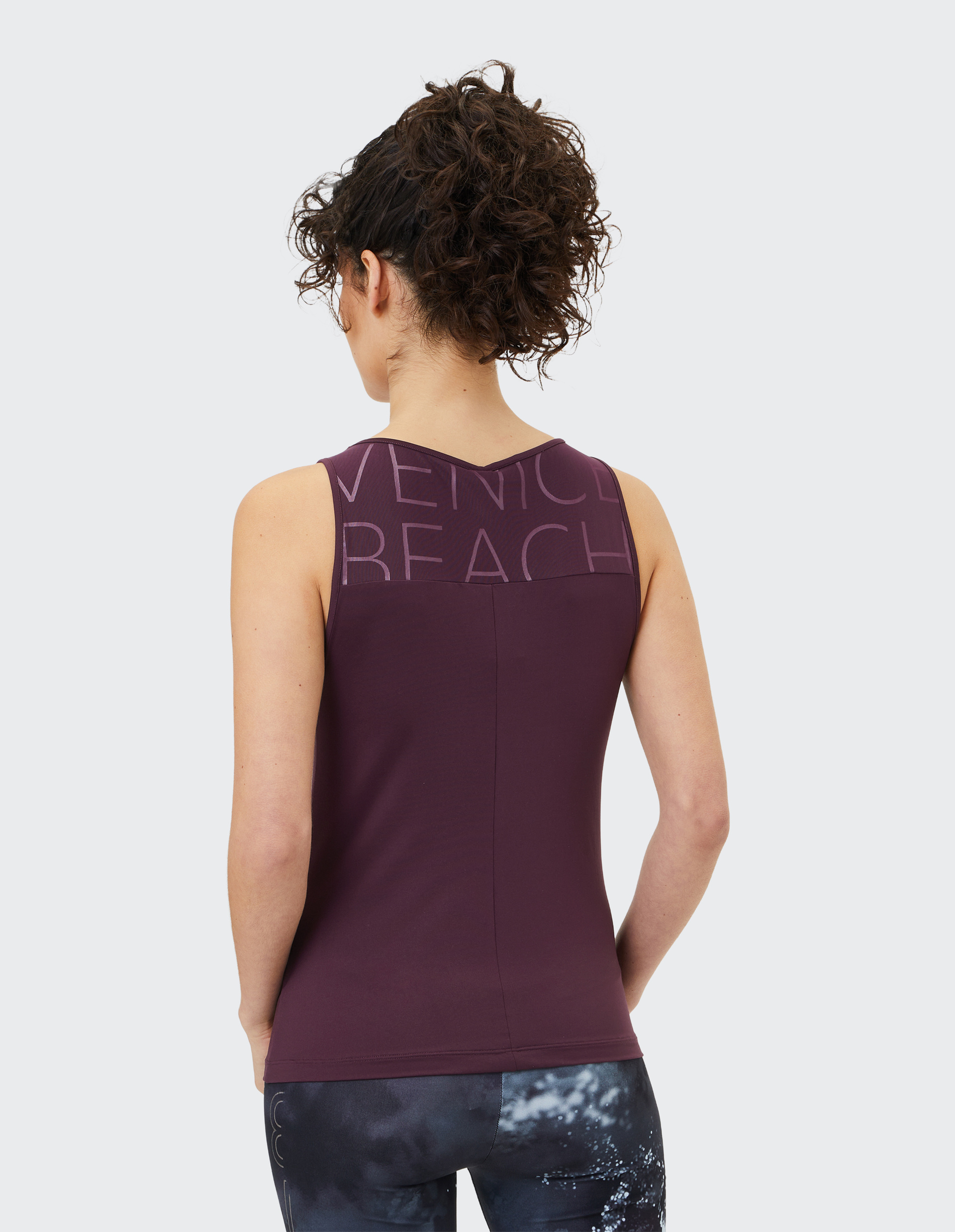 Frau mit lockigem Haar trägt ein ärmelloses, lila Oberteil mit Schriftzug und dunkle Leggings mit hellem, wolkenartigem Muster.