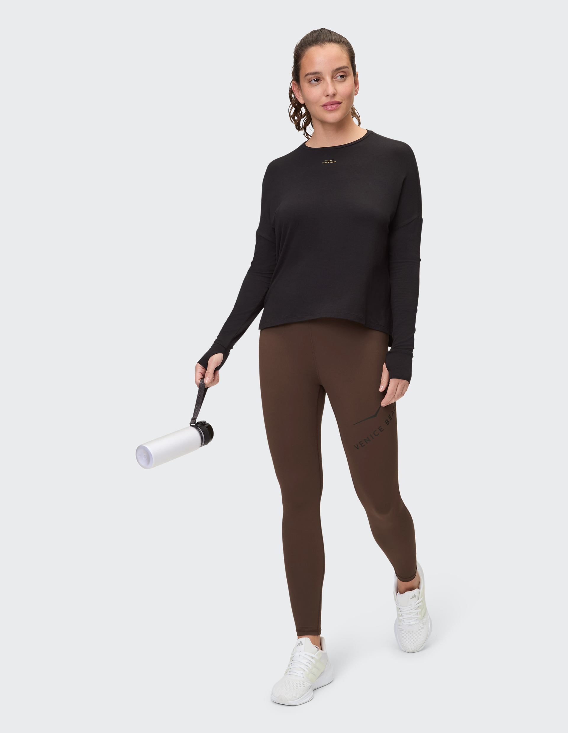Frau trägt schwarzes Langarmshirt und braune Leggings, steht aufrecht vor hellgrauem Hintergrund, weiße Sneaker an den Füßen.