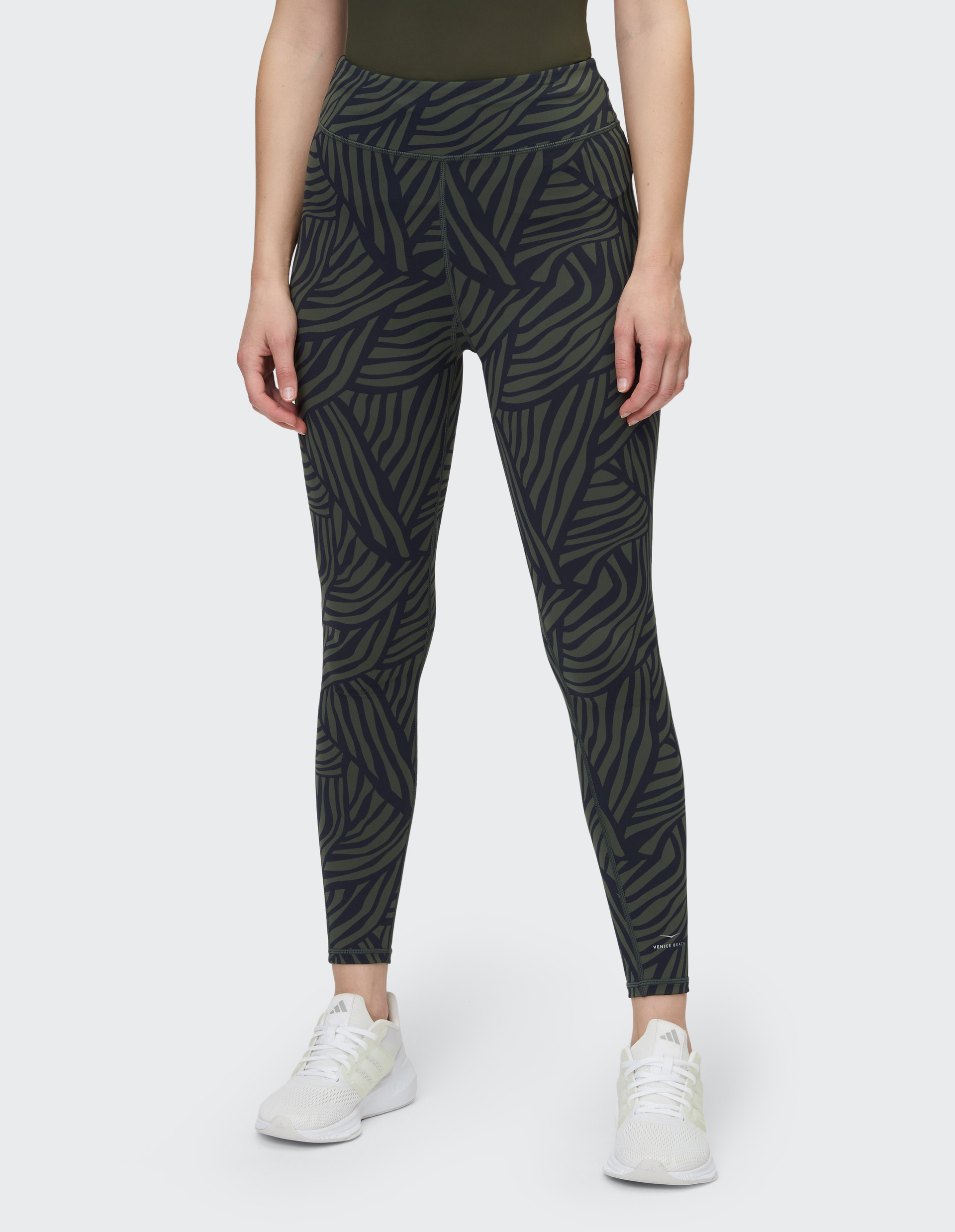 Frau trägt gemusterte Leggings und weiße Sneaker, steht aufrecht, zeigt die Beine und den unteren Oberkörper auf grauem Hintergrund.