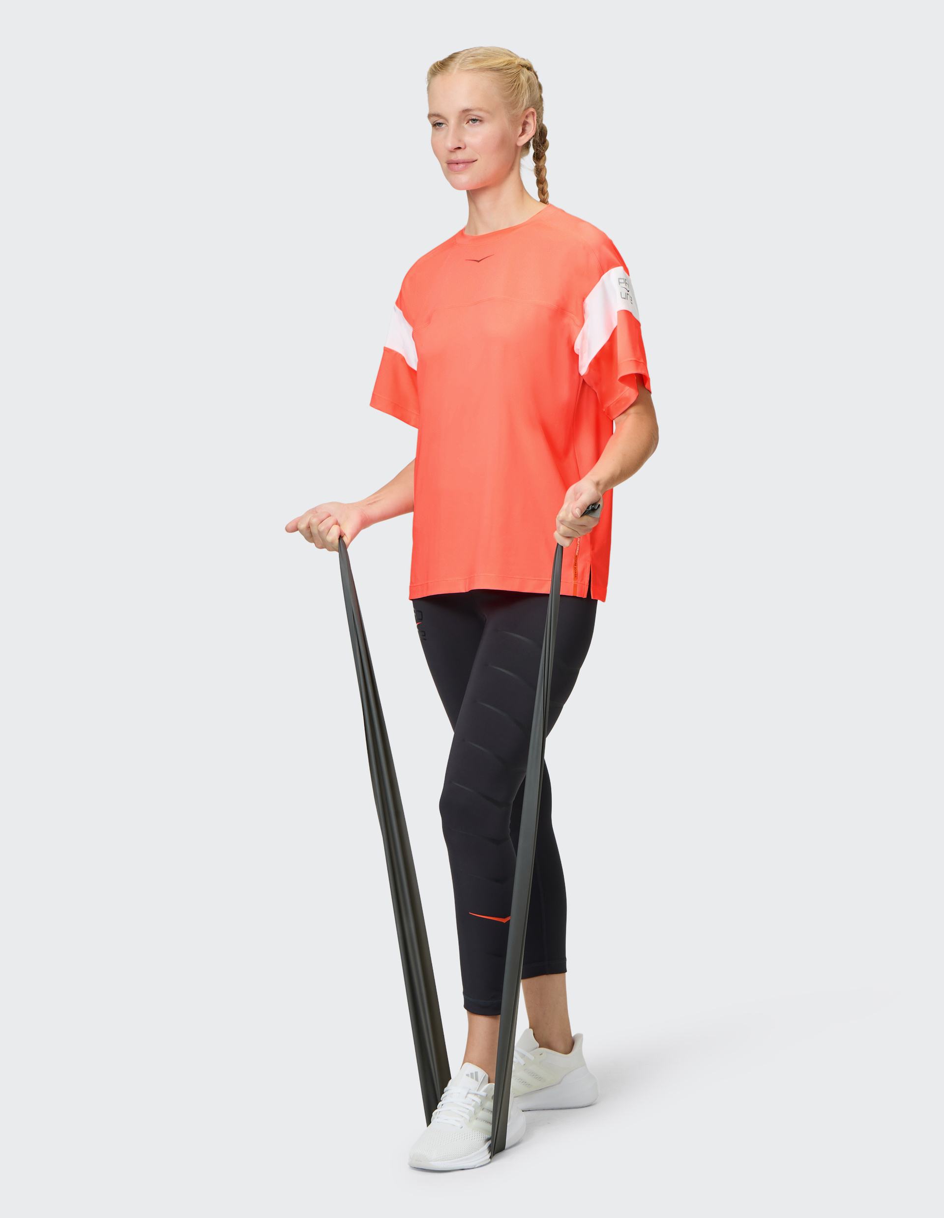 Frau in rotem Shirt und schwarzen Leggings nutzt Fitnessband, trägt weiße Sneakers, blondes Haar in Zopf, neutrale Miene.