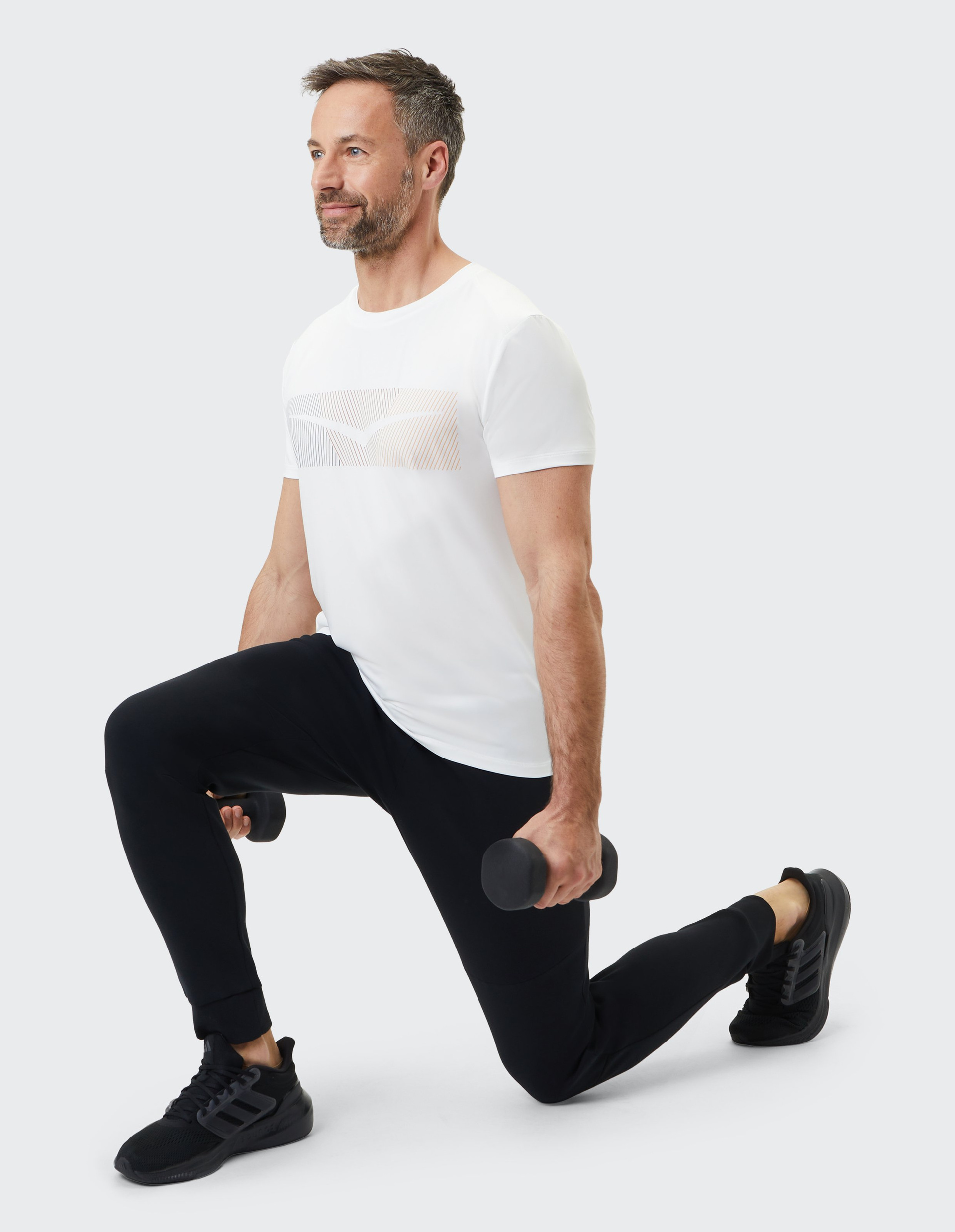 Ein Mann im weißen T-Shirt macht einen Ausfallschritt mit einer Hantel in jeder Hand, auf hellem Hintergrund.