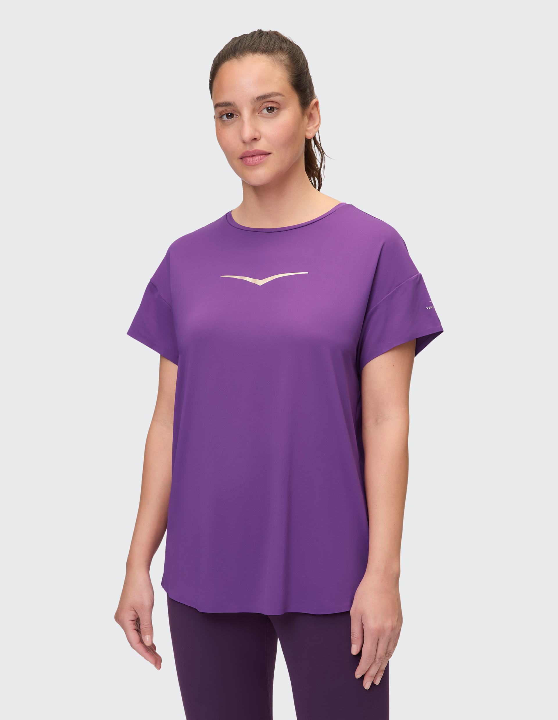 Frau in lila Sportkleidung steht vor neutralem Hintergrund; trägt lila T-Shirt mit Logo und passende Leggings.