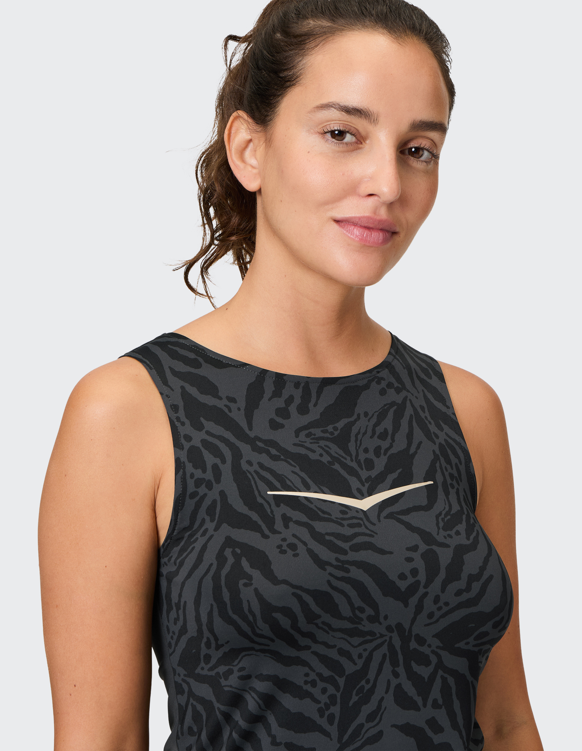 Eine Frau mit lockigem Haar trägt ein ärmelloses, schwarzes Top mit dunklem, abstraktem Muster und einem dezenten Lächeln.