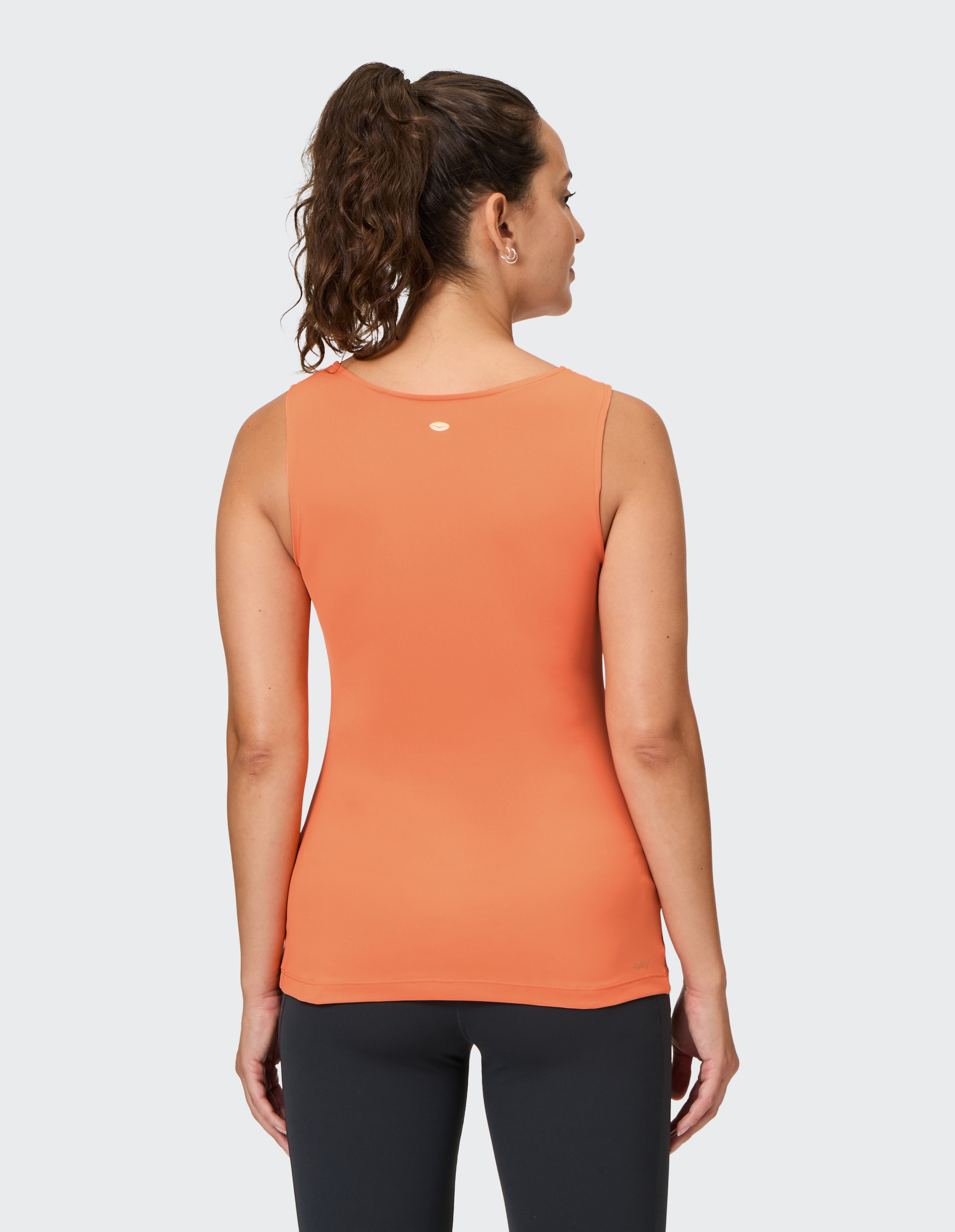 Frau mit lockigem Pferdeschwanz trägt ein orangefarbenes ärmelloses Top und schwarze Leggings, schaut über die Schulter auf grauem Hintergrund.