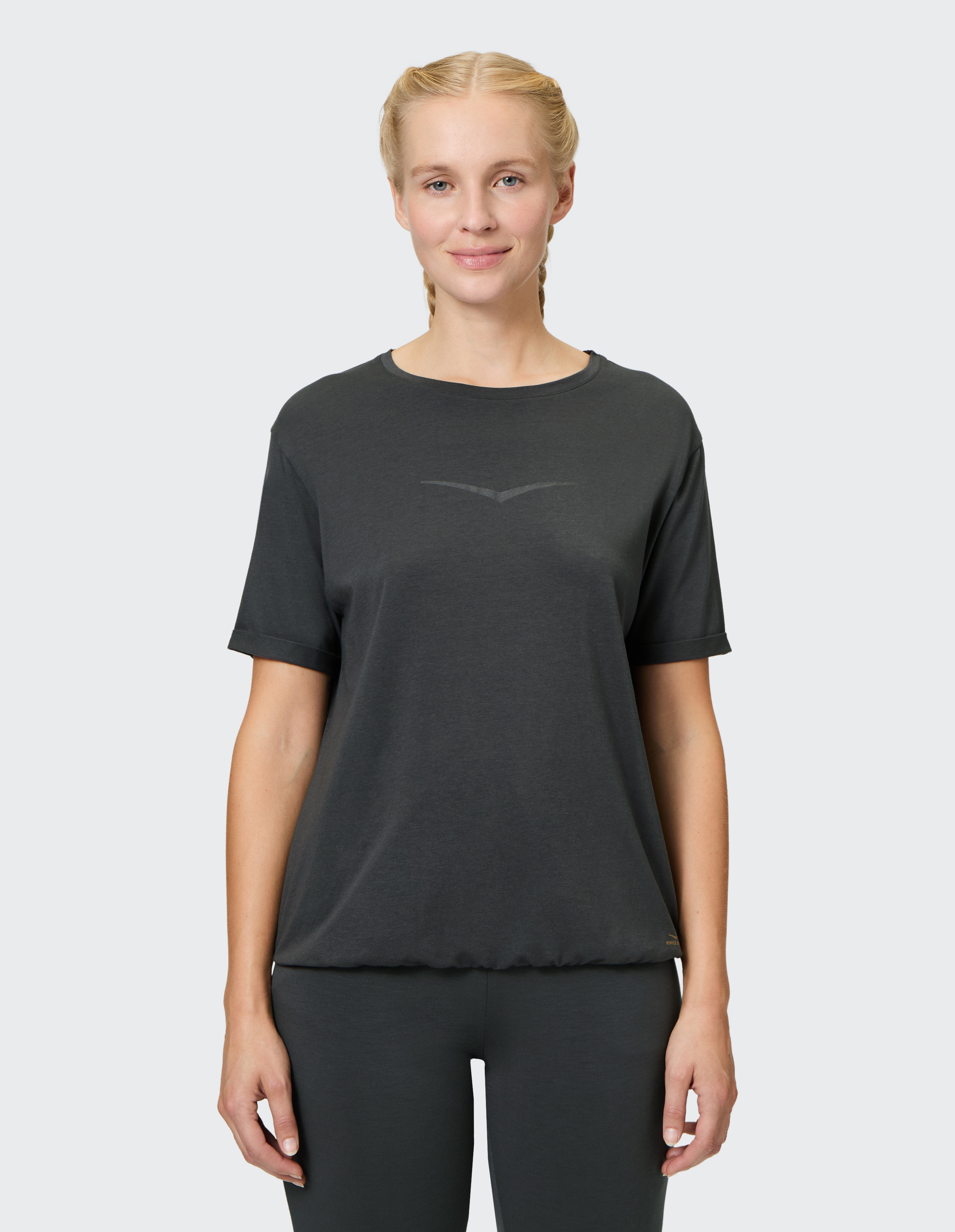 Eine Person in einem dunkelgrauen T-Shirt und Leggings steht frontal vor einem hellgrauen Hintergrund und lächelt leicht.