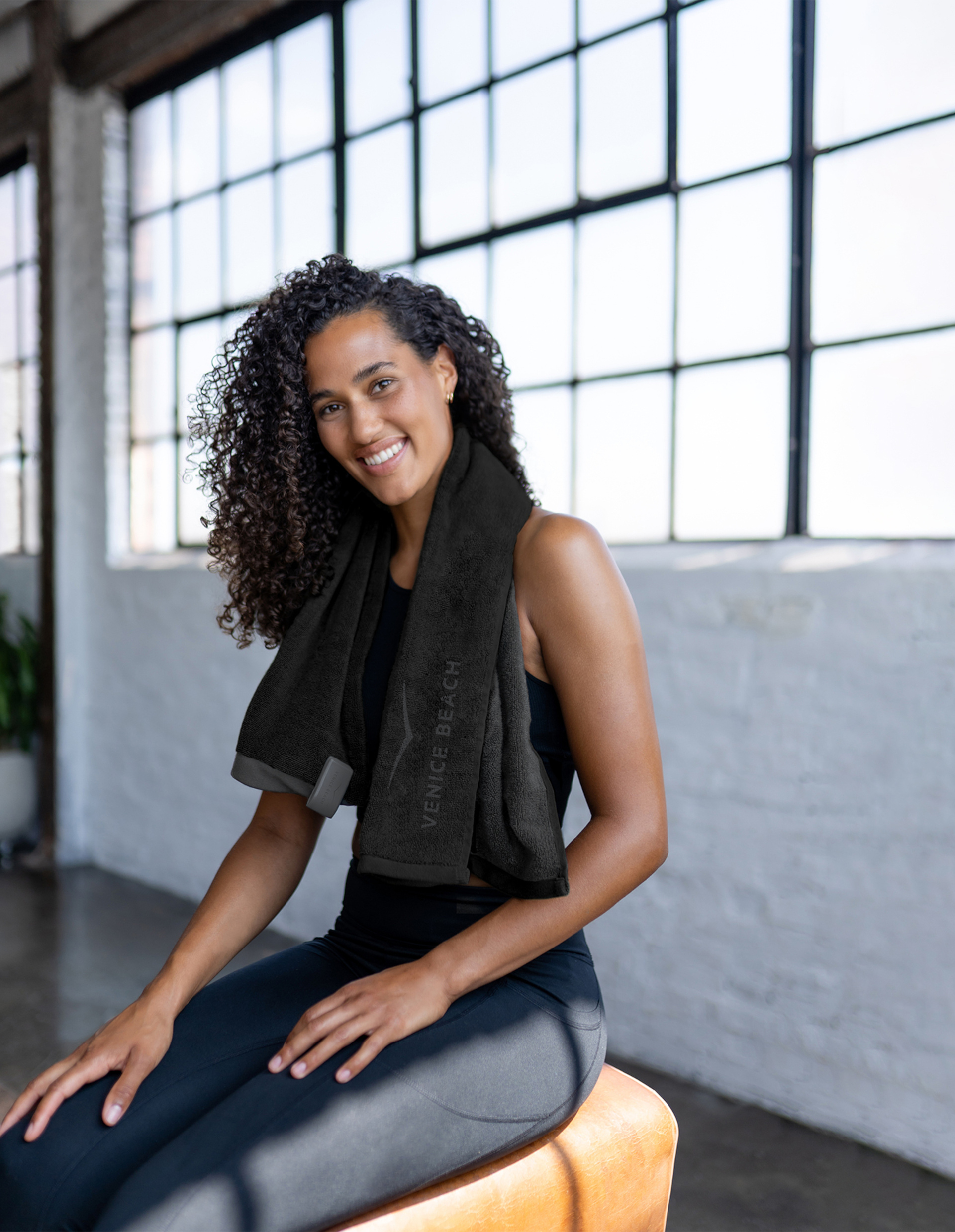 Frau in schwarzer Sportkleidung sitzt lächelnd mit Handtuch um den Nacken auf Lederhocker in hell erleuchtetem Raum mit großen Fenstern.