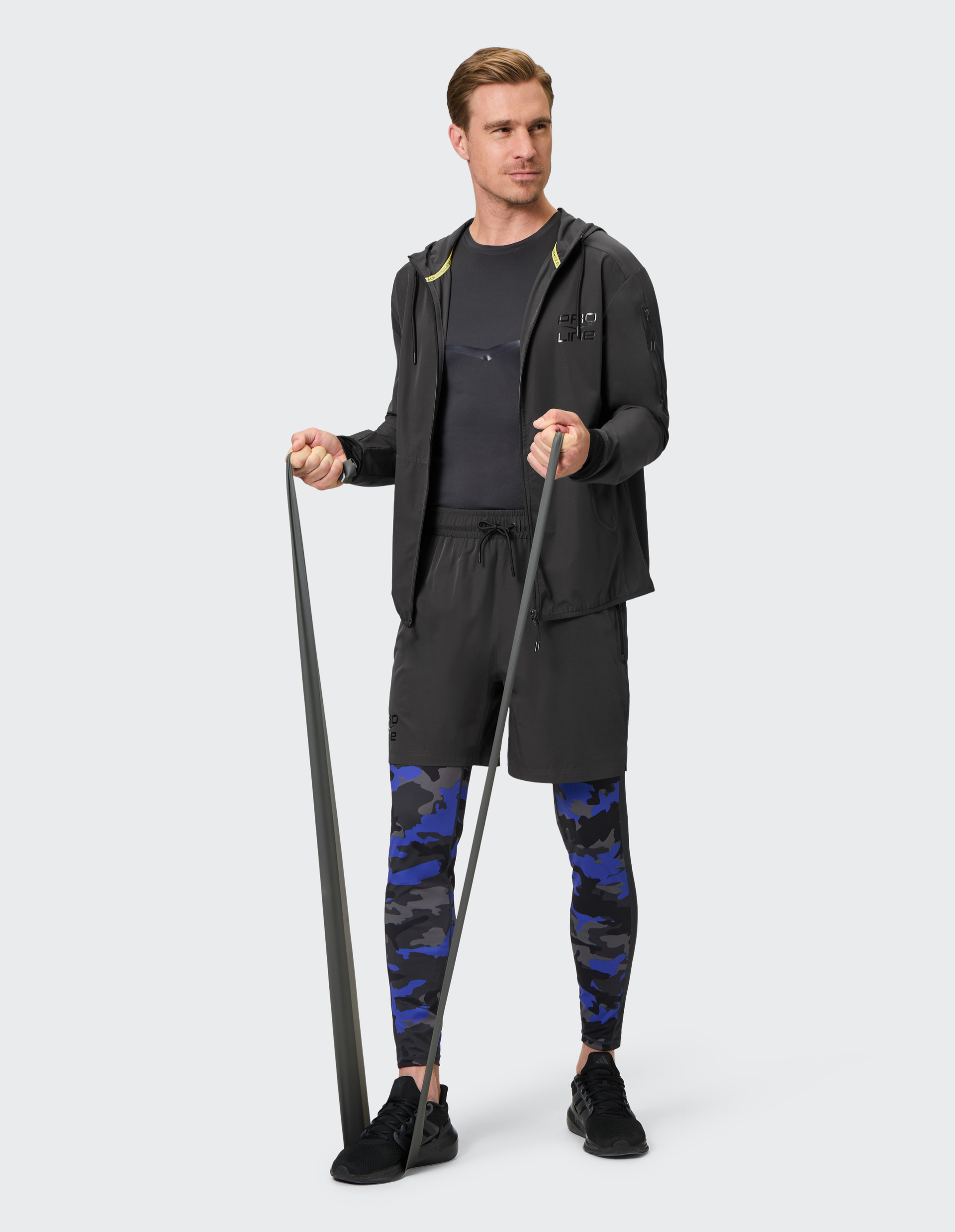 Ein Mann trainiert mit einem Fitnessband, trägt eine schwarze Sportjacke, kurze Hose und blau-camouflage Leggings, vor grauem Hintergrund.
