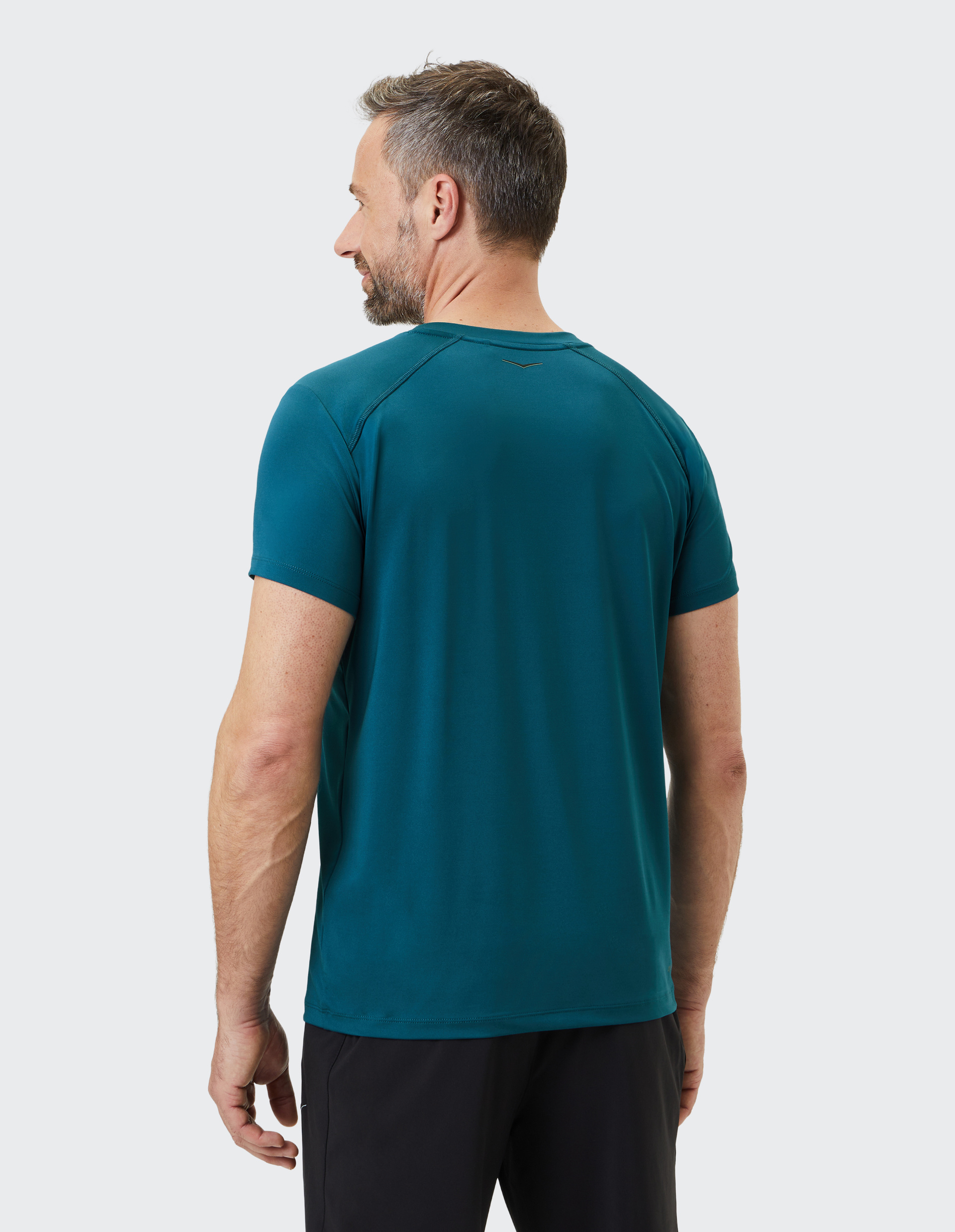 Ein Mann mit kurzem Haar in einem türkisen T-Shirt und schwarzen Shorts steht mit dem Rücken zur Kamera vor einem hellgrauen Hintergrund.