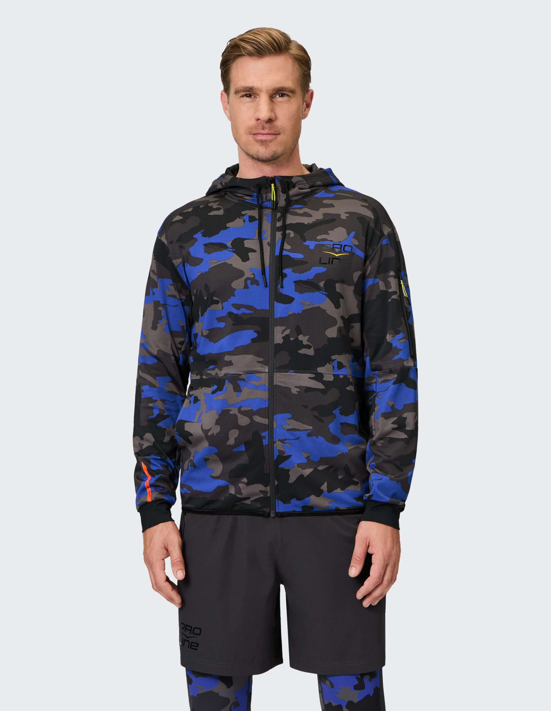Ein Mann trägt eine blau-schwarz-graue Camouflage-Jacke mit Kapuze, kombiniert mit dunklen Shorts und gemusterten Leggings.