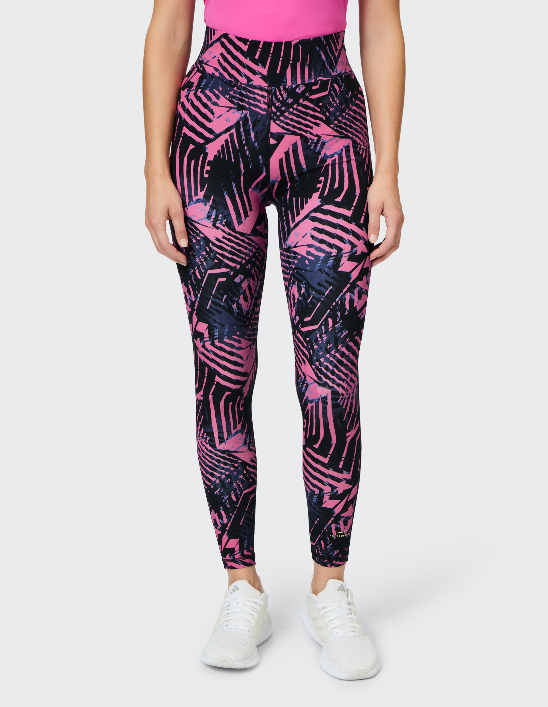 Frau trägt bunte, geometrisch gemusterte Leggings in Pink und Blau mit weißen Turnschuhen und einem pinken Oberteil.