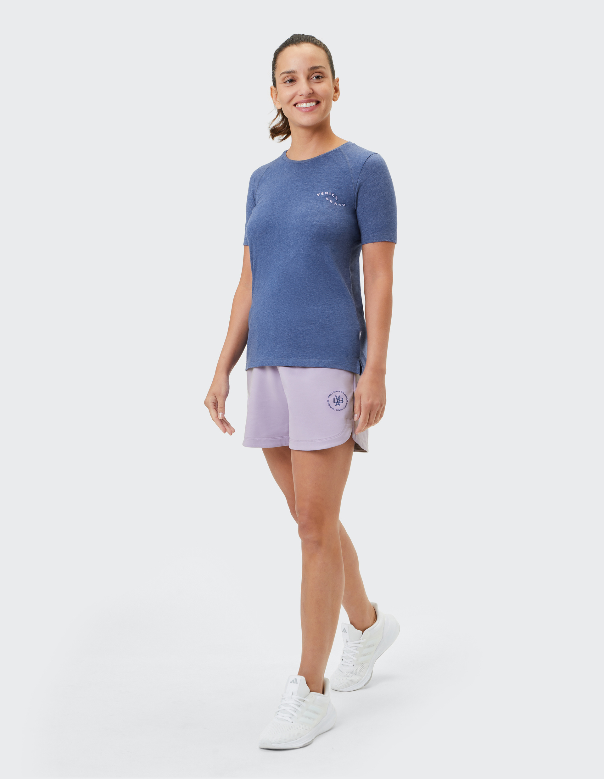 Frau in blauem T-Shirt und lila Shorts lächelt, geht nach links, trägt weiße Sneaker, hat lockeren Pferdeschwanz.