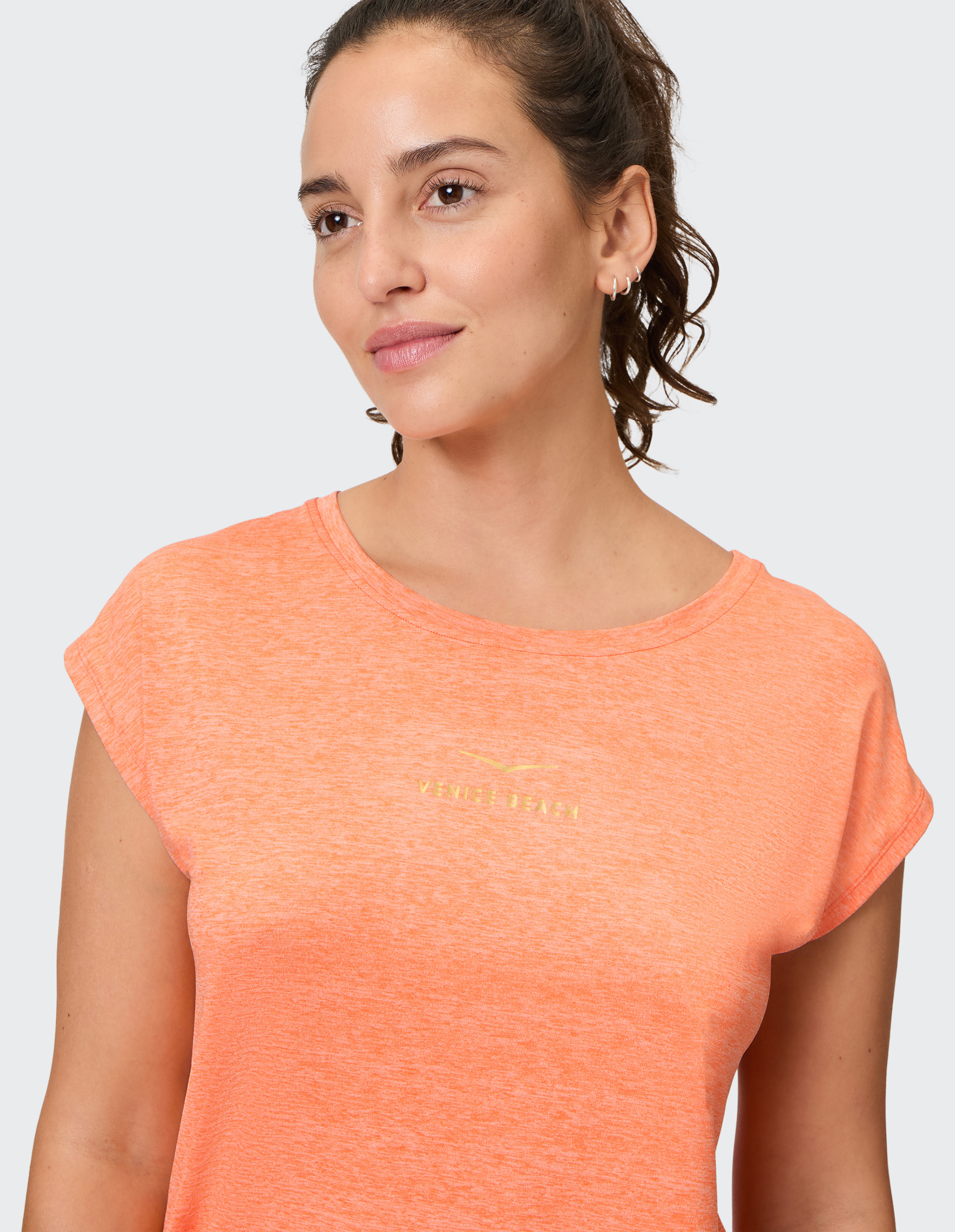 Frau mit lockigem Haar trägt ein orangefarbenes T-Shirt mit goldenen Buchstaben auf der Brust und blickt nach rechts.