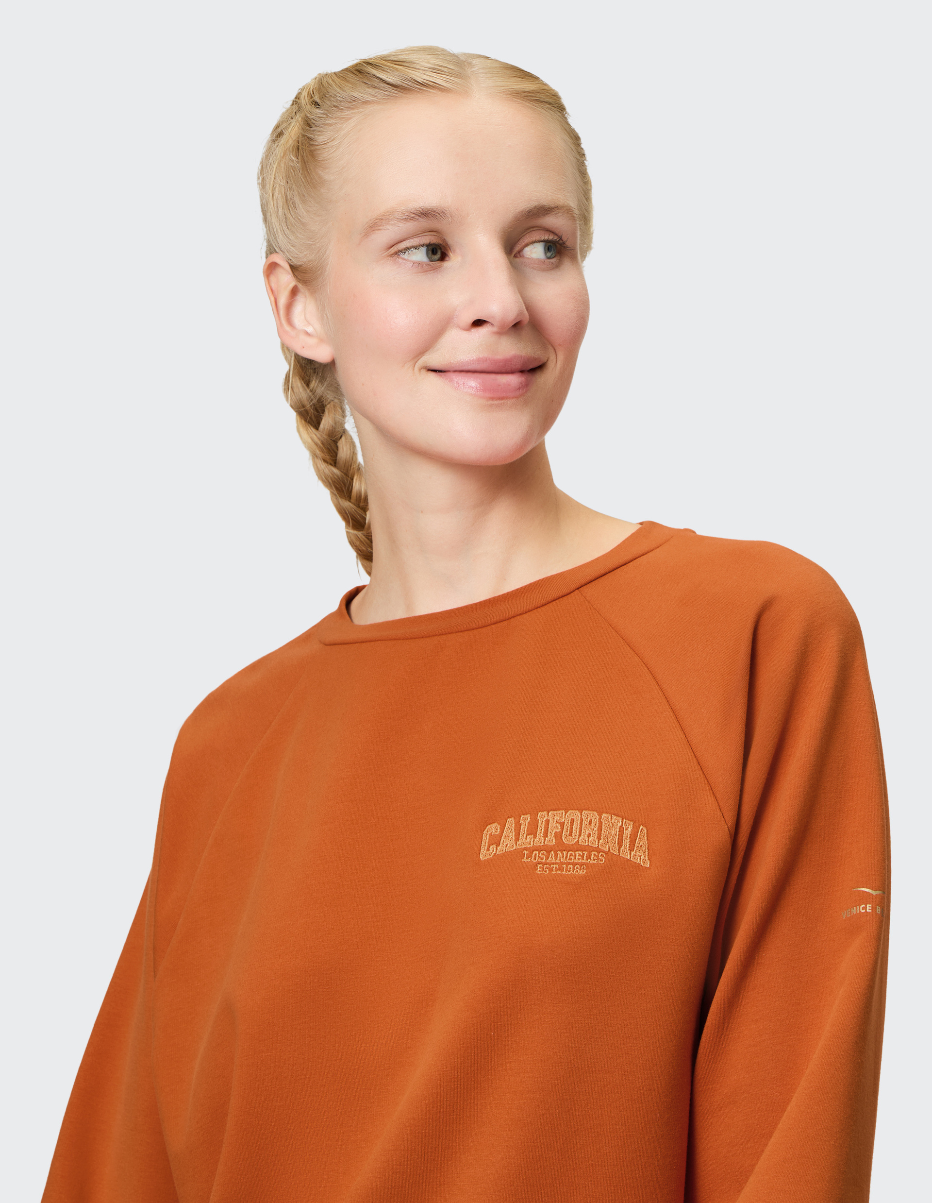 Eine lächelnde Frau mit blonden Zöpfen trägt ein orangefarbenes Sweatshirt mit "California" Aufdruck und blickt nach rechts.