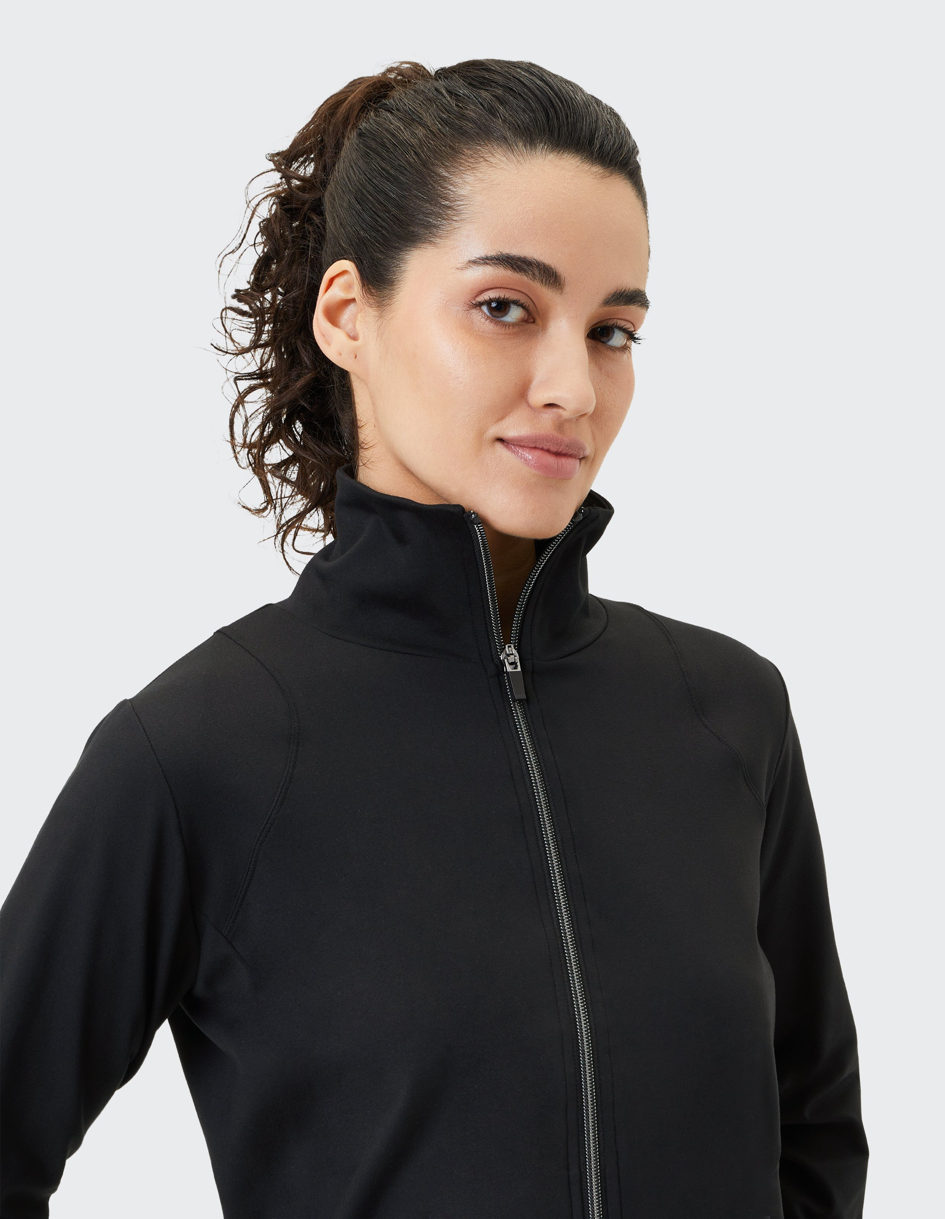 Frau mit lockigem Pferdeschwanz trägt hochgeschlossene schwarze Jacke mit Reißverschluss vor neutralem Hintergrund.