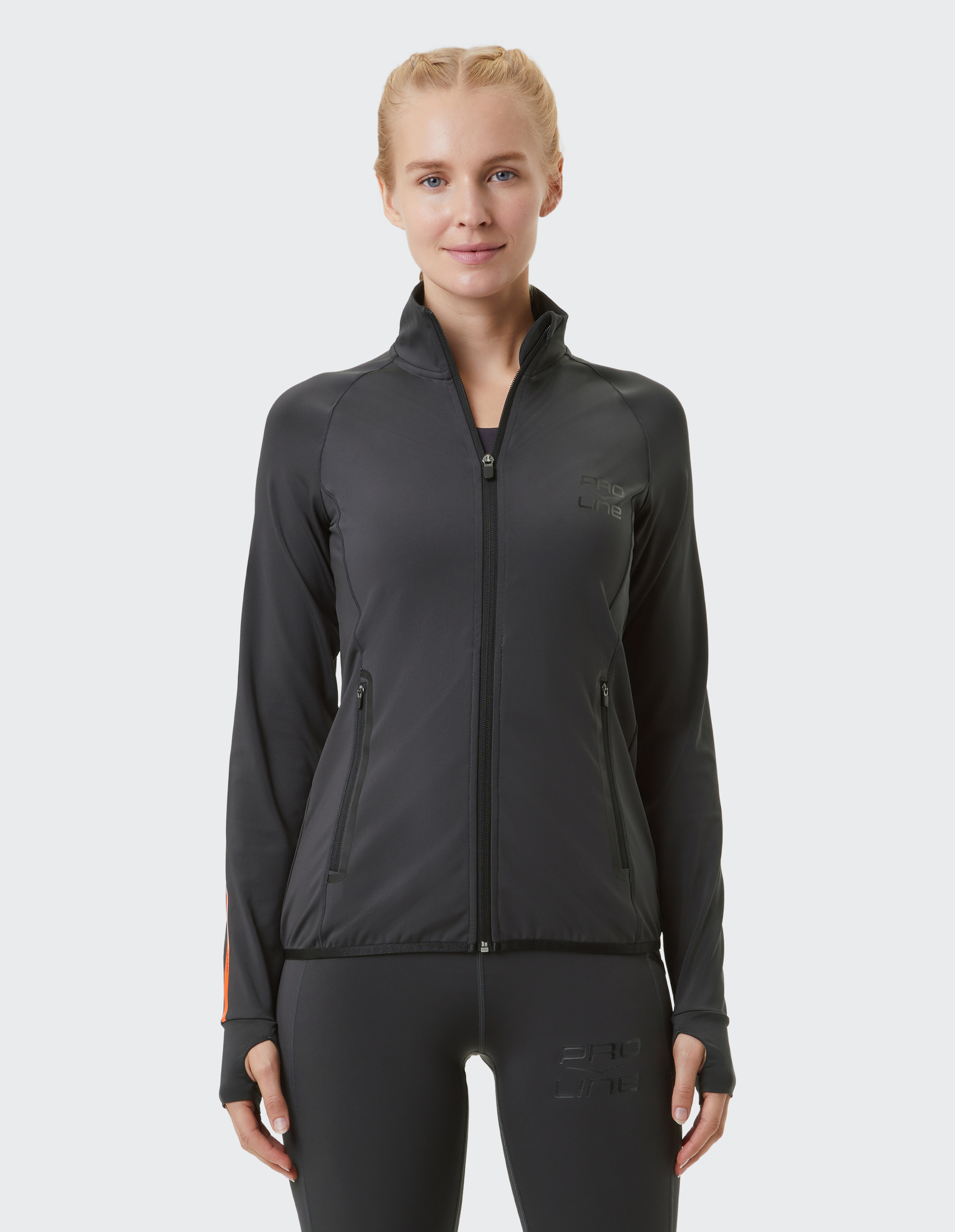 Frau in schwarzer Sportjacke mit Reißverschluss und schlichter Hose, lächelt leicht, vor hellem Hintergrund.