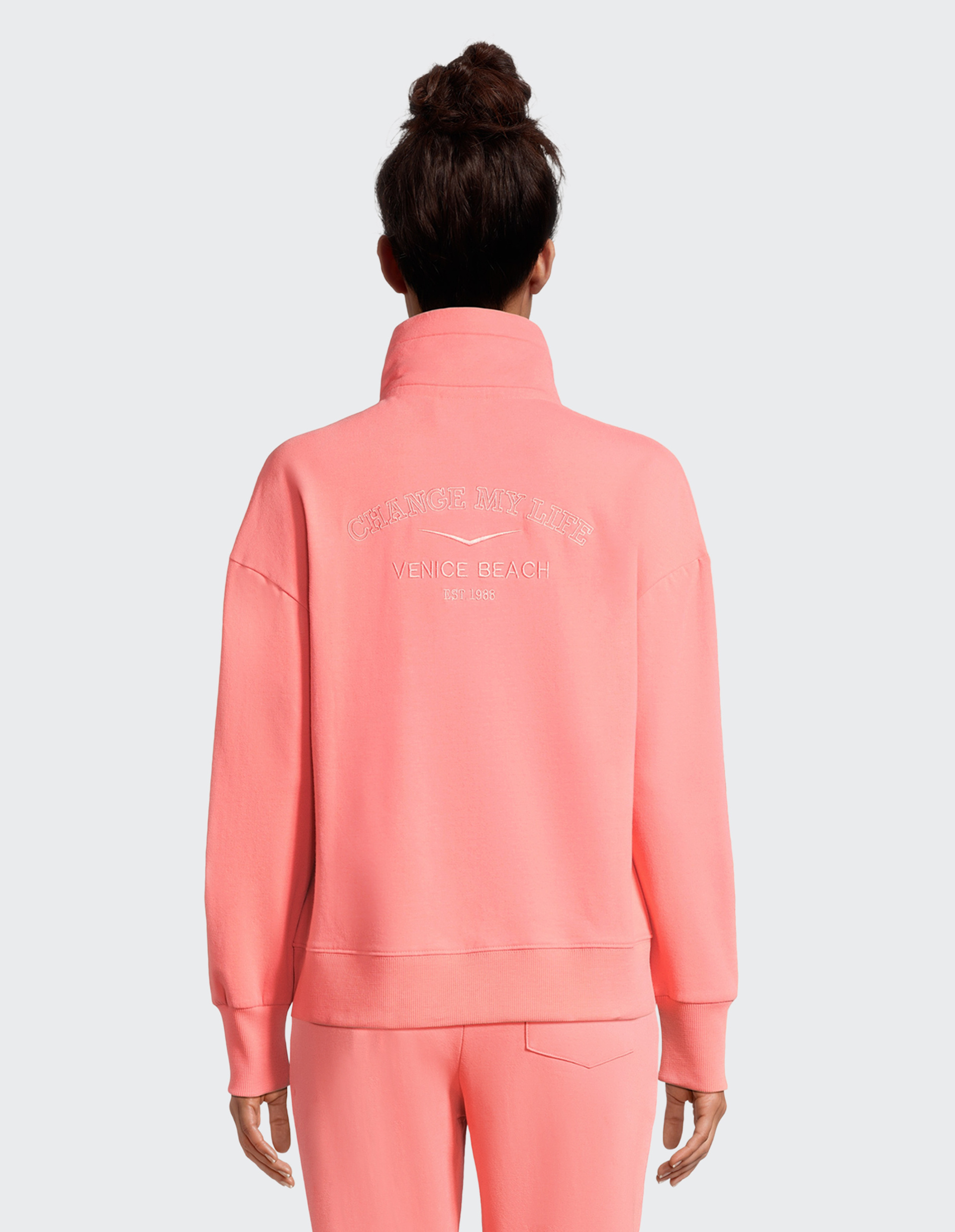 Person in rosa Trainingsanzug mit hochgestecktem Haar, Rückenaufschrift auf dem Pullover: "CHANGE MY LIFE VENICE BEACH EST 1986".