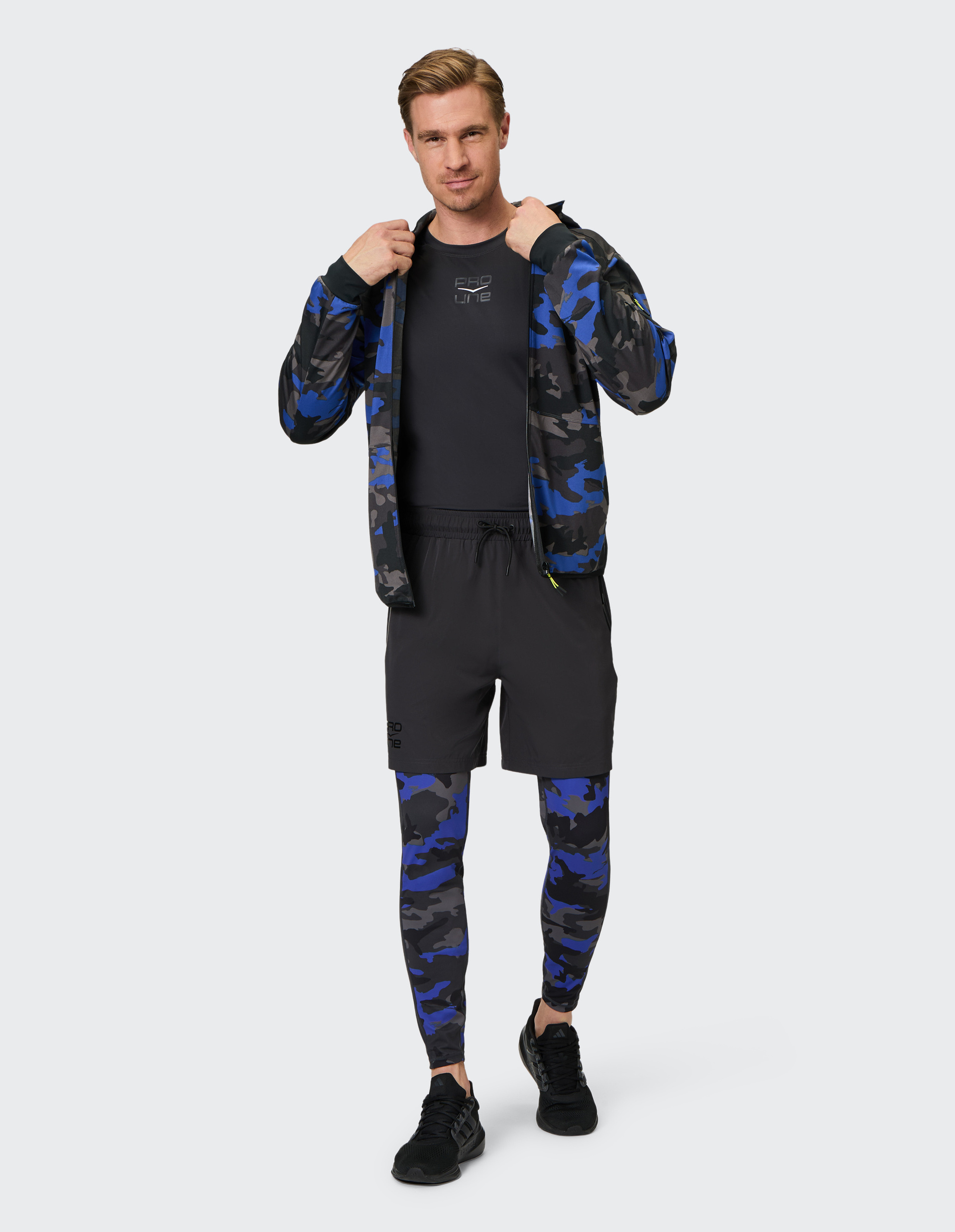 Ein Mann trägt eine blau-schwarz-graue Tarnjacke mit passenden Leggings, ein schwarzes Shirt, Shorts und Sneakers.