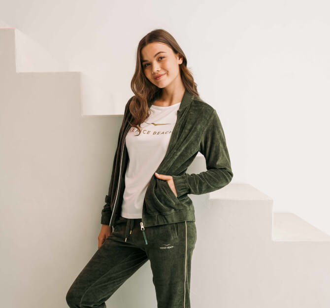 Frau in dunkelgrünem Jogginganzug und weißem T-Shirt mit goldenem Venice Beach Logo.