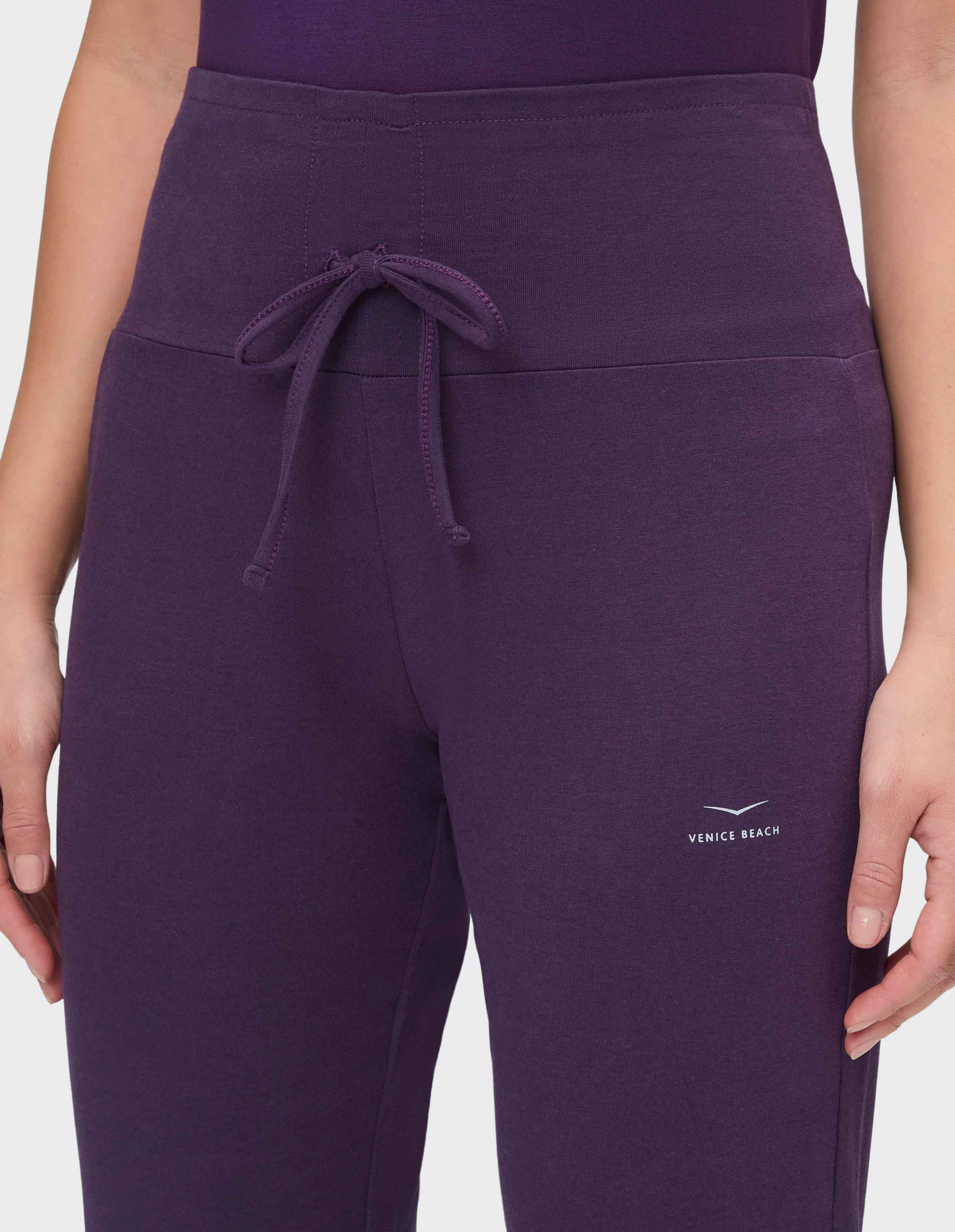 Frau trägt lila Jogginghose mit breitem Bund und Zierkordel, Logo "Venice Beach" über Knie, Arme locker an den Seiten.