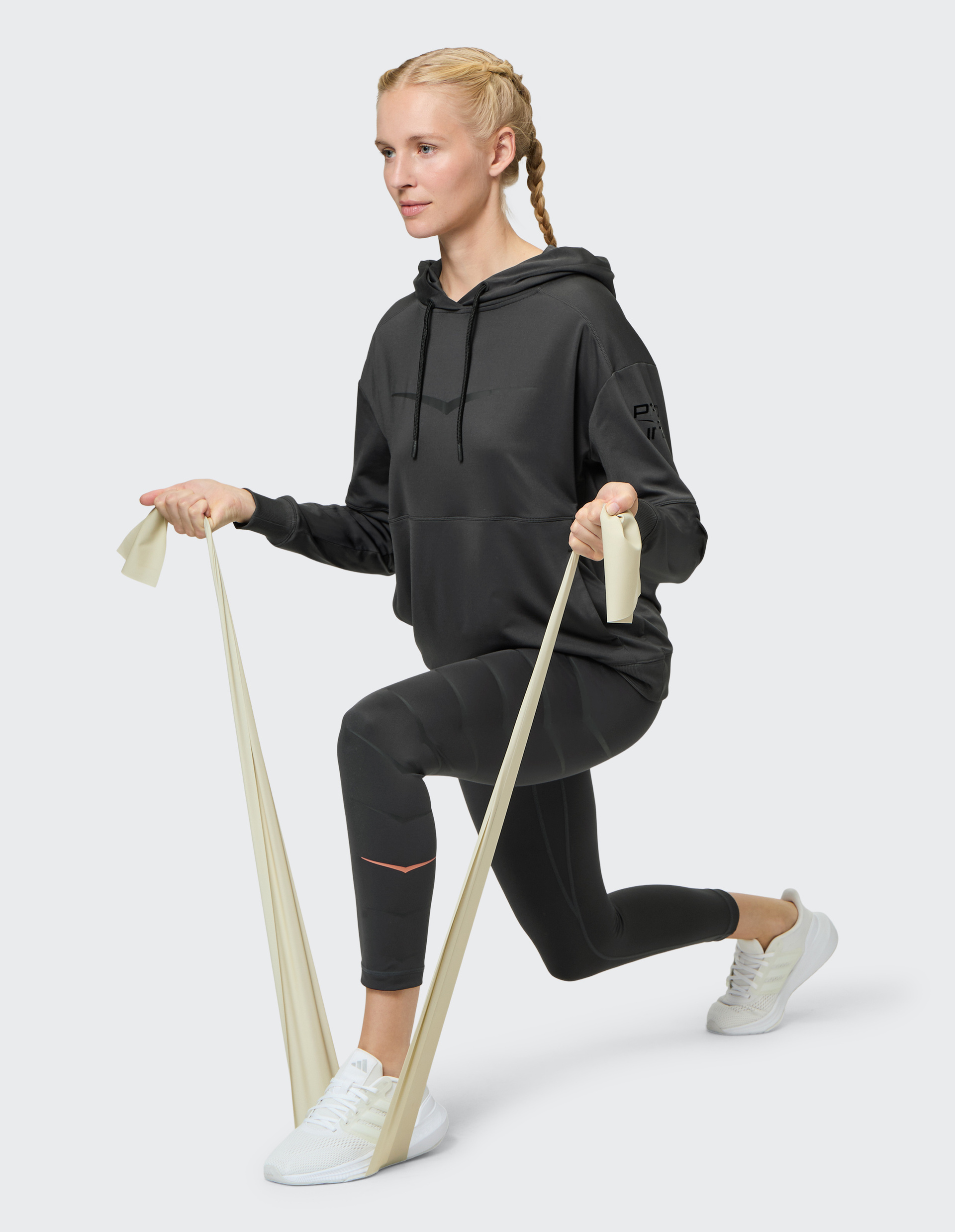Eine Person mit geflochtenem Haar macht einen Ausfallschritt und nutzt ein Widerstandsband, trägt Sportkleidung und weiße Sneakers.