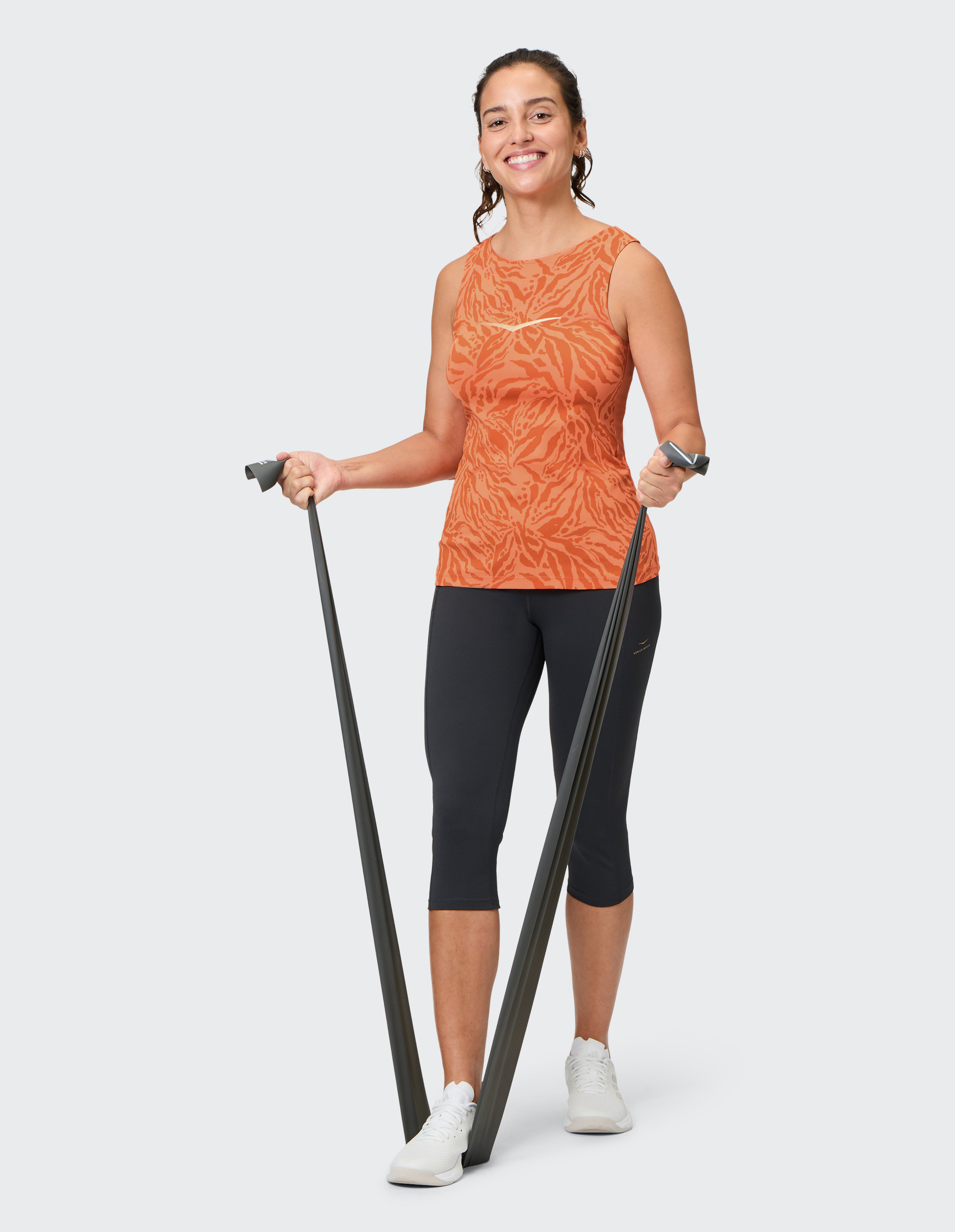 Frau in orangefarbenem Top und schwarzen Leggings trainiert mit Fitnessband, während sie lächelnd in die Kamera blickt.