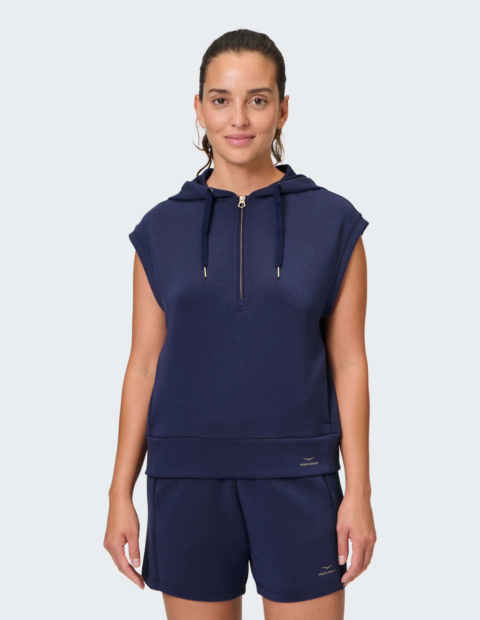 Frau in dunkelblauem ärmellosen Hoodie mit Reißverschluss und passenden Shorts, stehend vor einem neutralen Hintergrund.