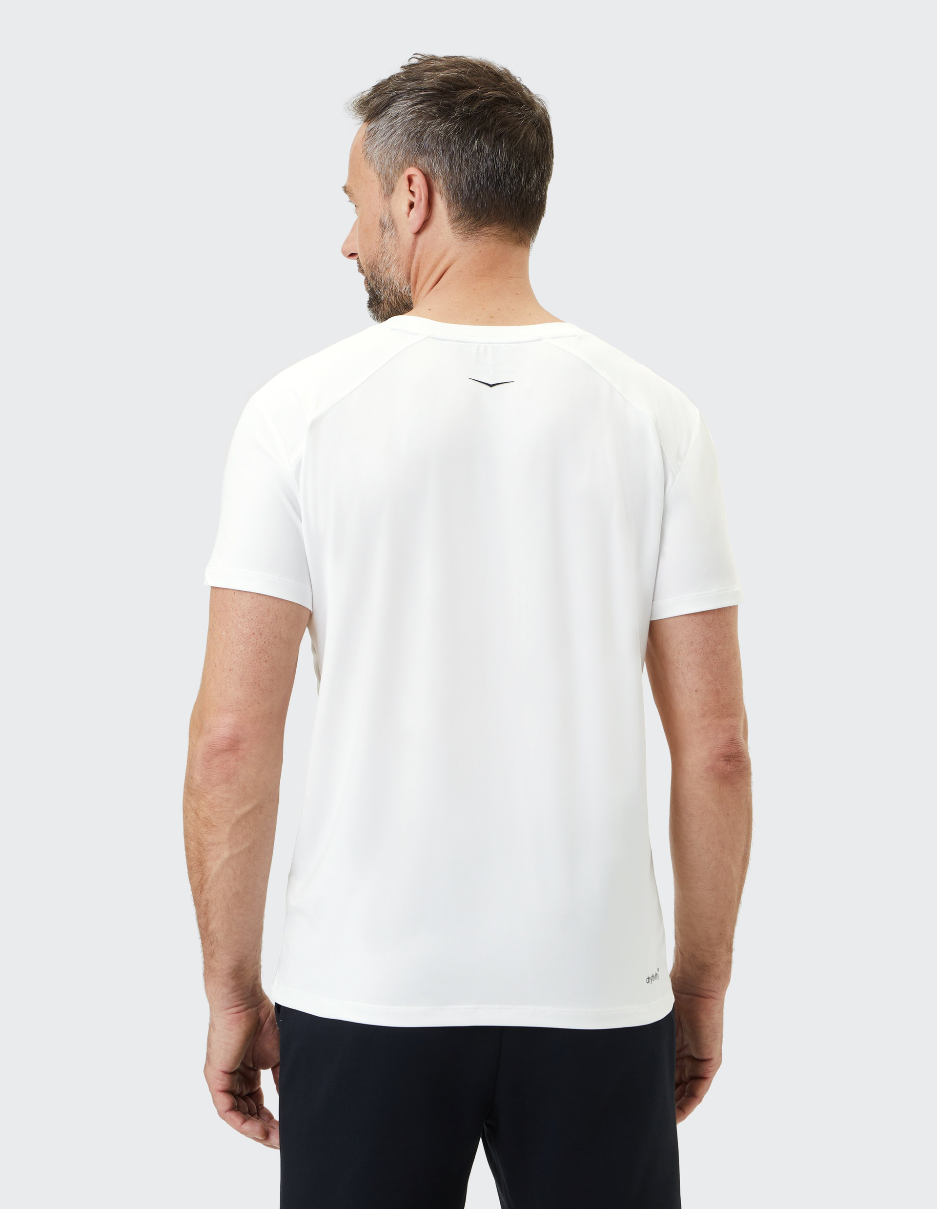 Ein Mann mit kurzem Haar trägt ein weißes T-Shirt und schwarze Hose, von hinten gesehen, vor einem hellgrauen Hintergrund.