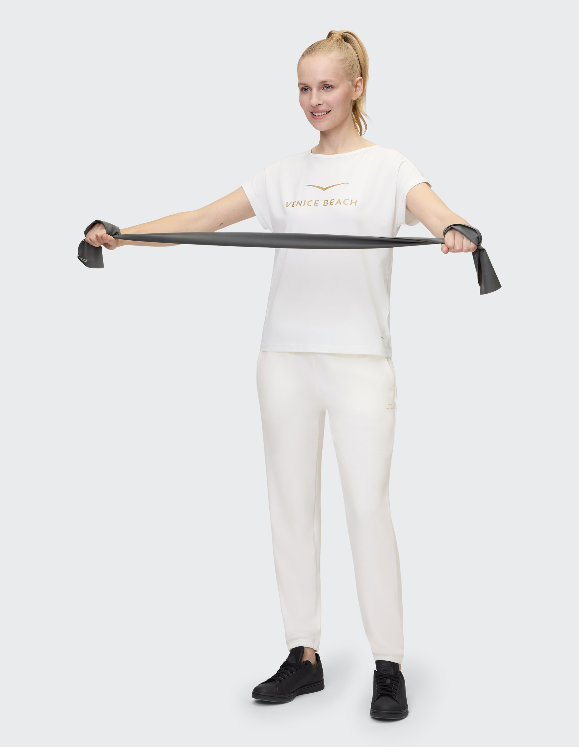 Person in einem weißen "Venice Beach"-Shirt und Hose, trägt schwarze Sneaker, steht vor einem hellgrauen Hintergrund.