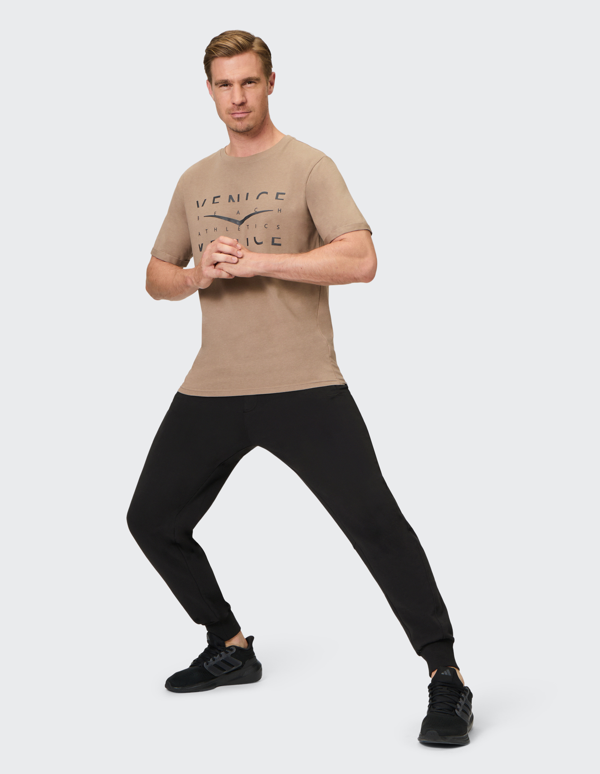 Ein Mann in beigefarbenem T-Shirt und schwarzer Hose steht in Kampfpose, die Hände sind vor dem Körper gefaltet.