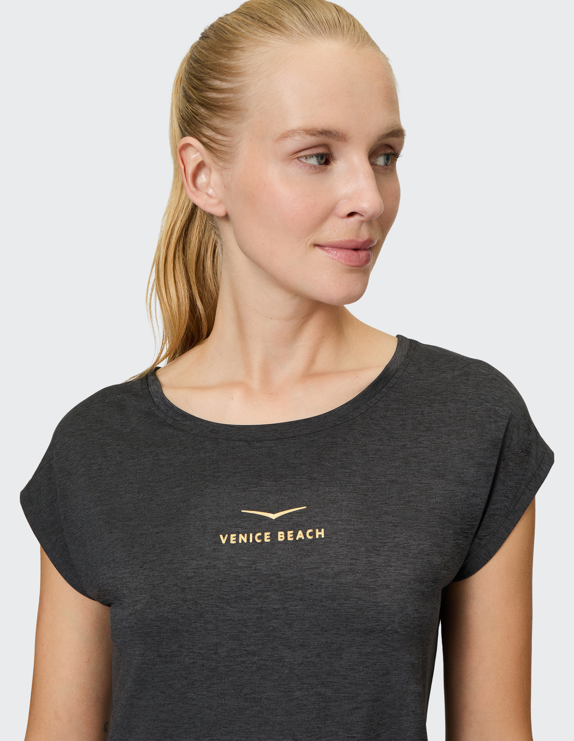 Frau mit blondem Pferdeschwanz trägt ein graues, kurzärmliges T-Shirt mit "Venice Beach"-Schriftzug und blickt zur Seite.