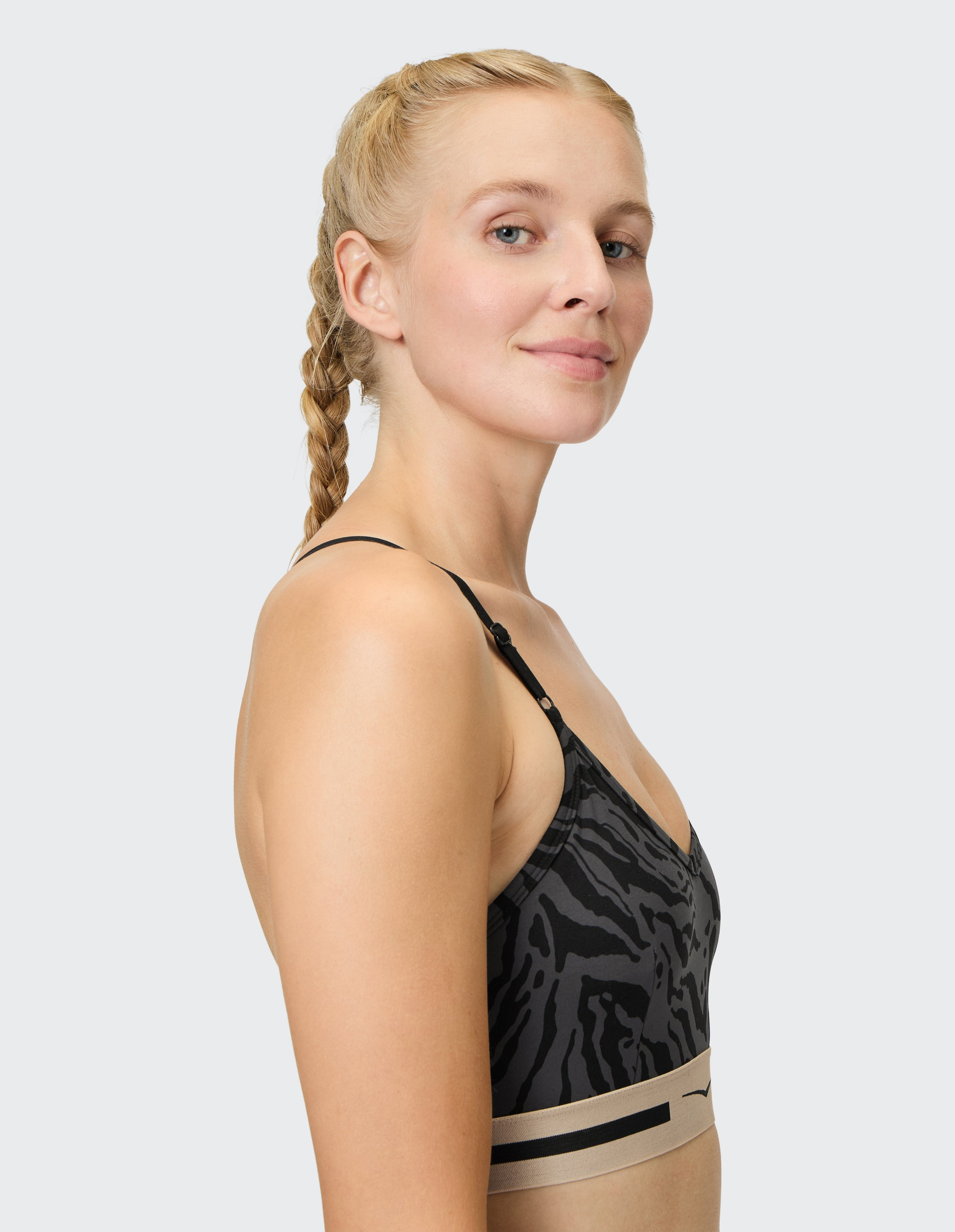 Blonde Frau im Seitprofil mit geflochtenem Haar trägt einen schwarzen Sport-BH mit Zebramuster und beigem Band, vor grauem Hintergrund.