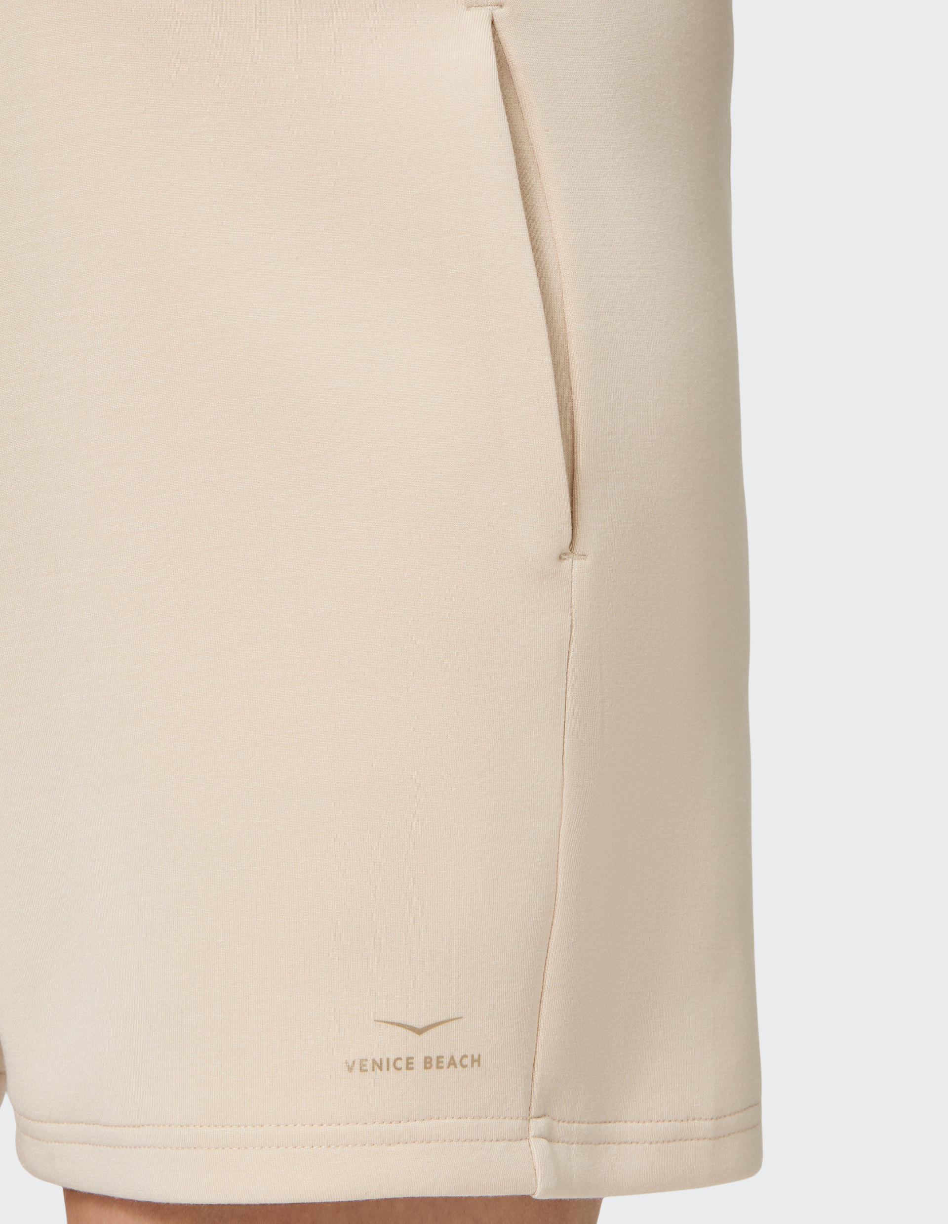 Nahaufnahme eines beigen Kleidungsstücks mit Taschen und dem Schriftzug "Venice Beach" am Saum auf hellem Hintergrund.