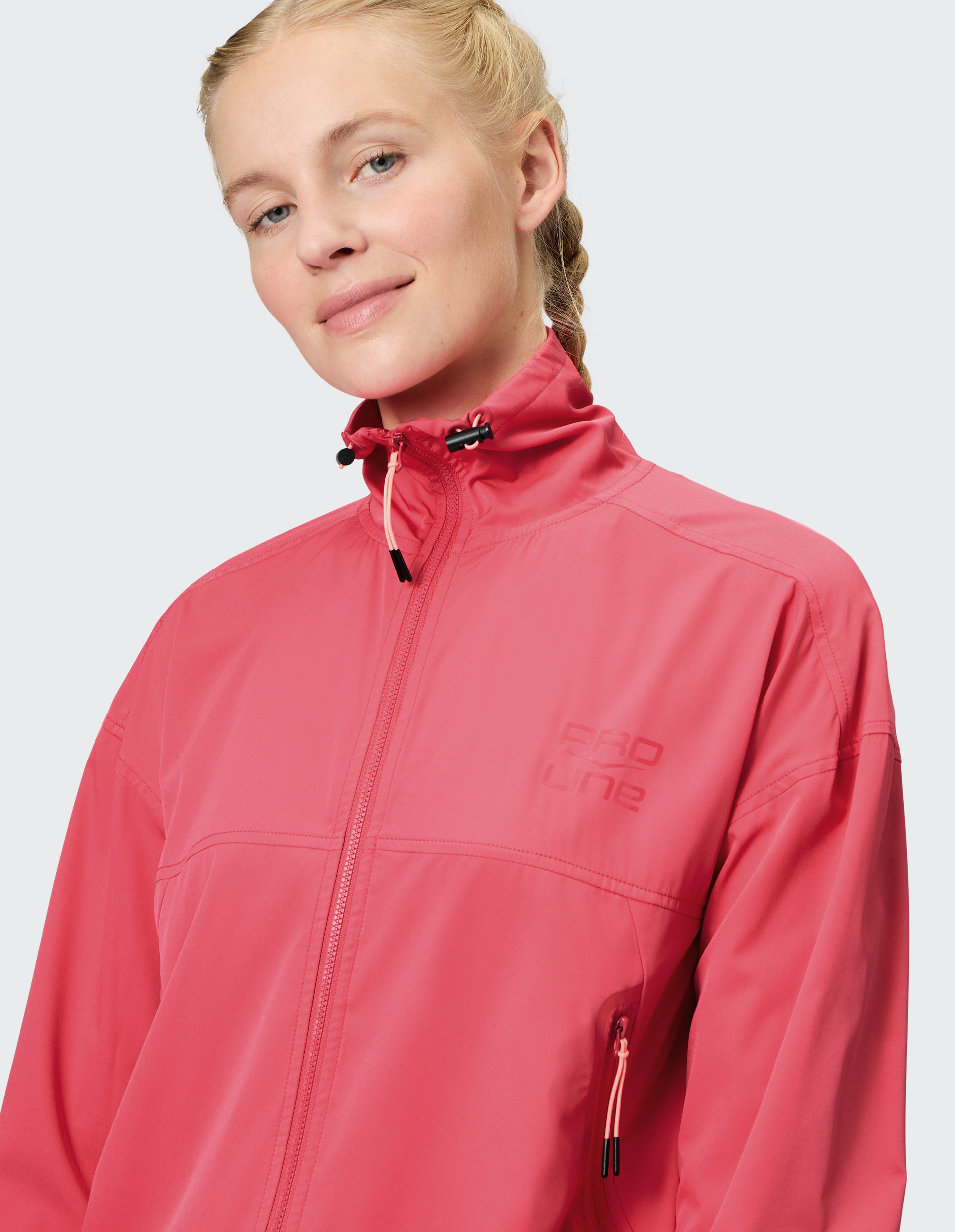 Eine lächelnde Person mit blonden Zöpfen trägt eine leuchtend rosa Jacke mit hohem Kragen und Reißverschluss vor grauem Hintergrund.