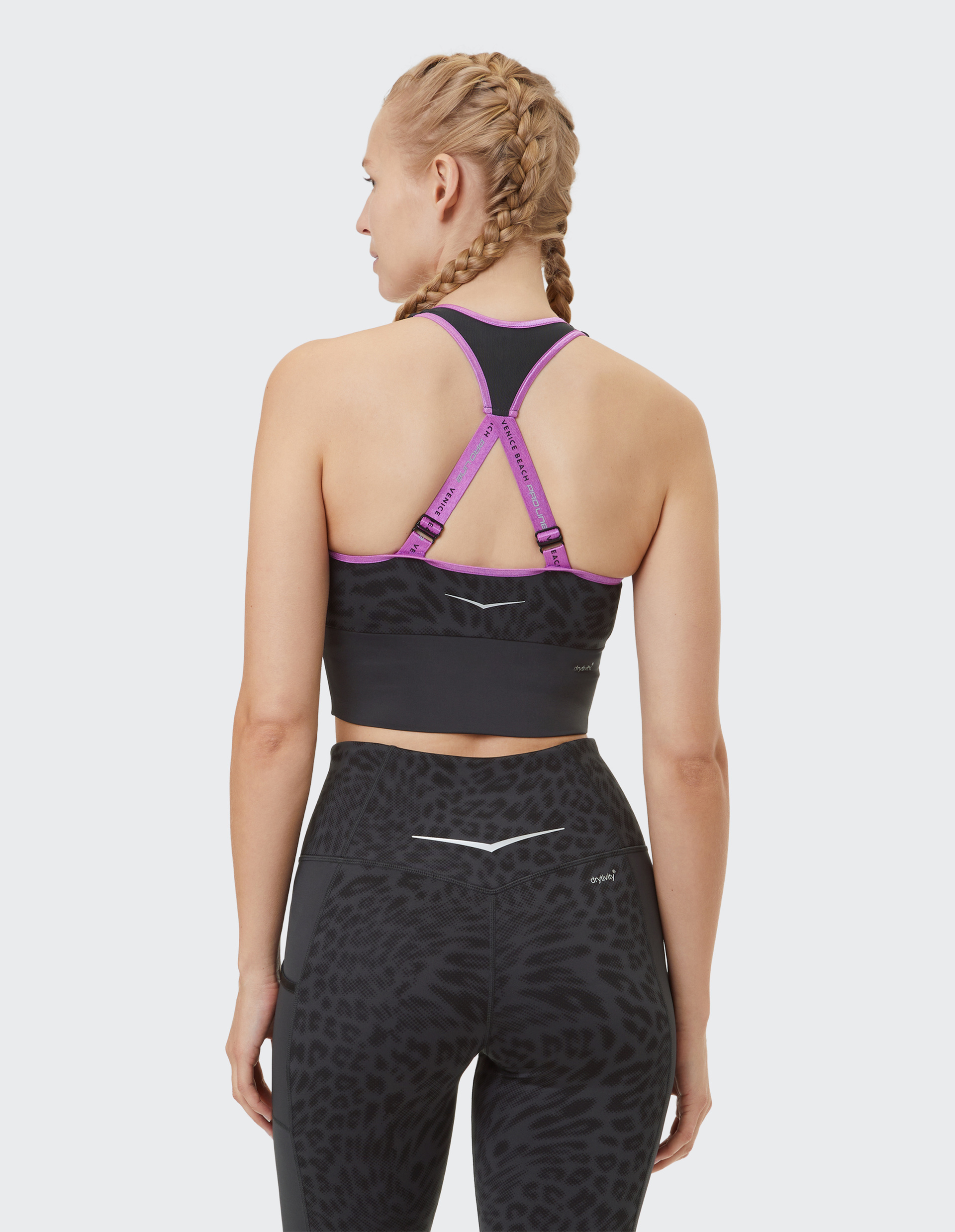 Eine Frau mit geflochtenem Haar trägt ein schwarzes und violettes Sporttop und schwarze Leggings mit Leopardenmuster, von hinten gesehen.