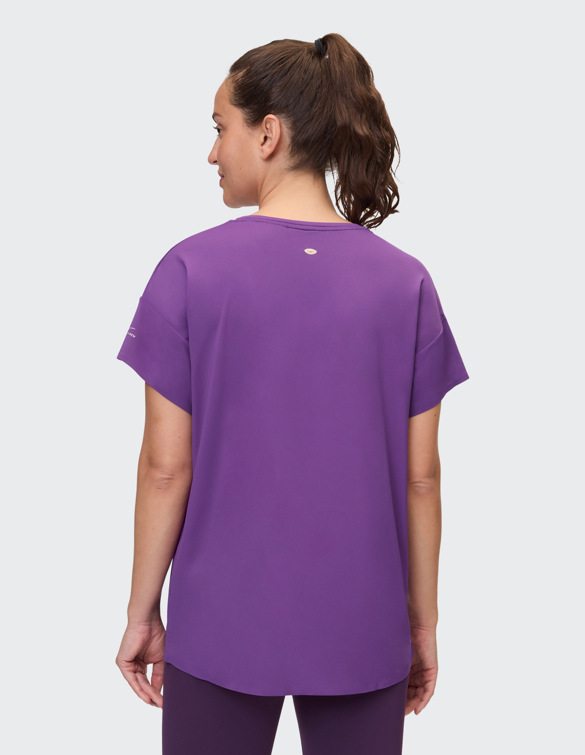 Frau mit Pferdeschwanz und lila T-Shirt steht seitlich und schaut nach links; Hintergrund ist neutral.