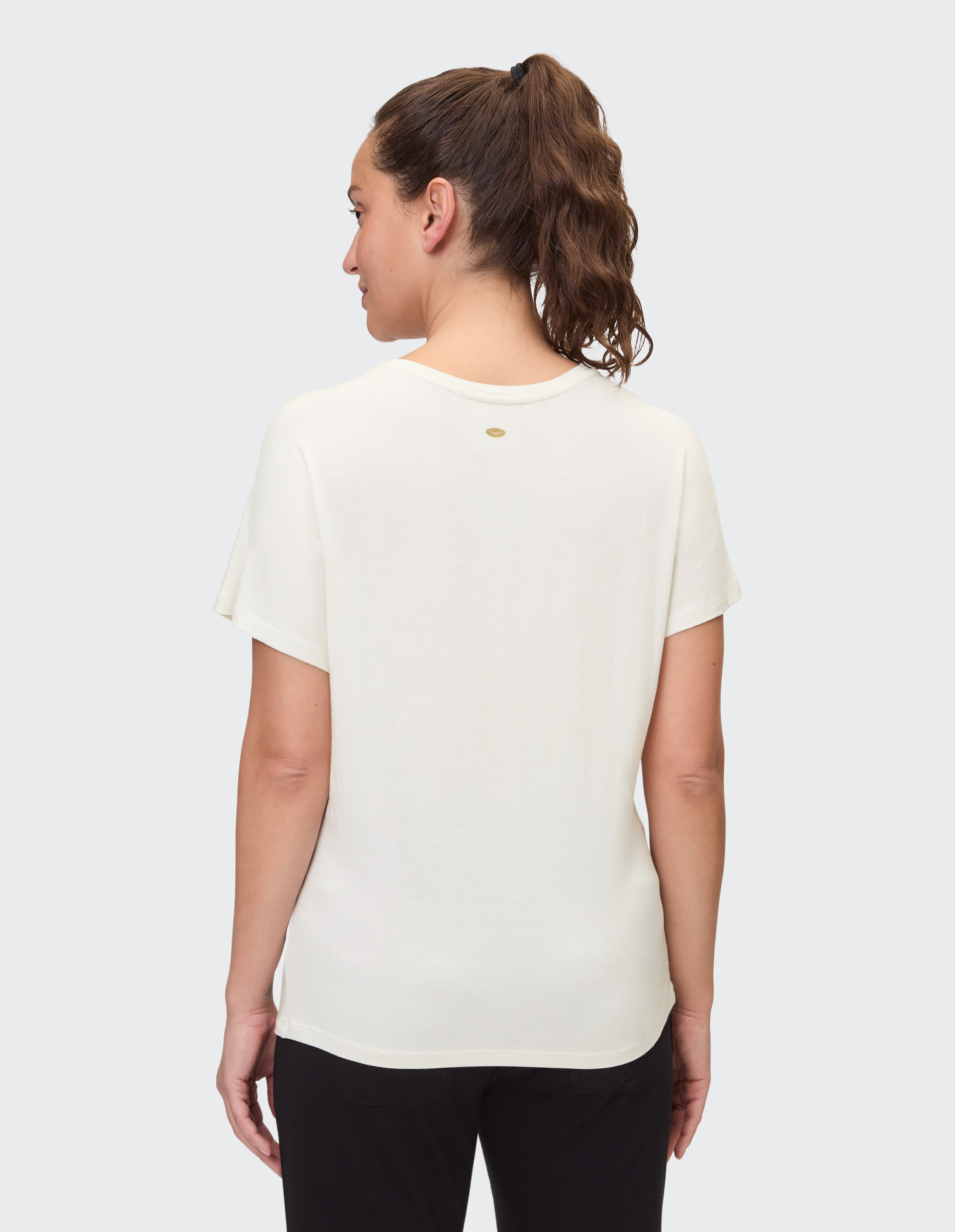 Eine Frau mit braunem Haar im Pferdeschwanz trägt ein cremeweißes T-Shirt und schwarze Hose, von hinten betrachtet.