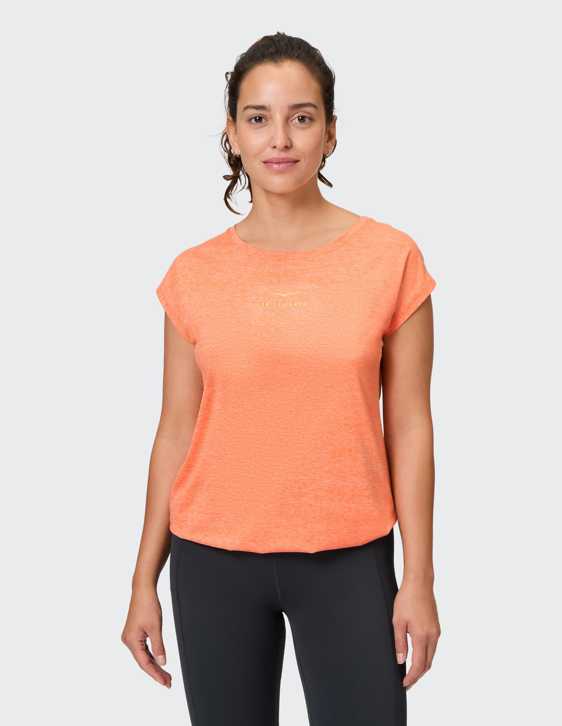 Frau in orangefarbenem T-Shirt und dunkler Hose lächelt leicht vor neutralem Hintergrund.