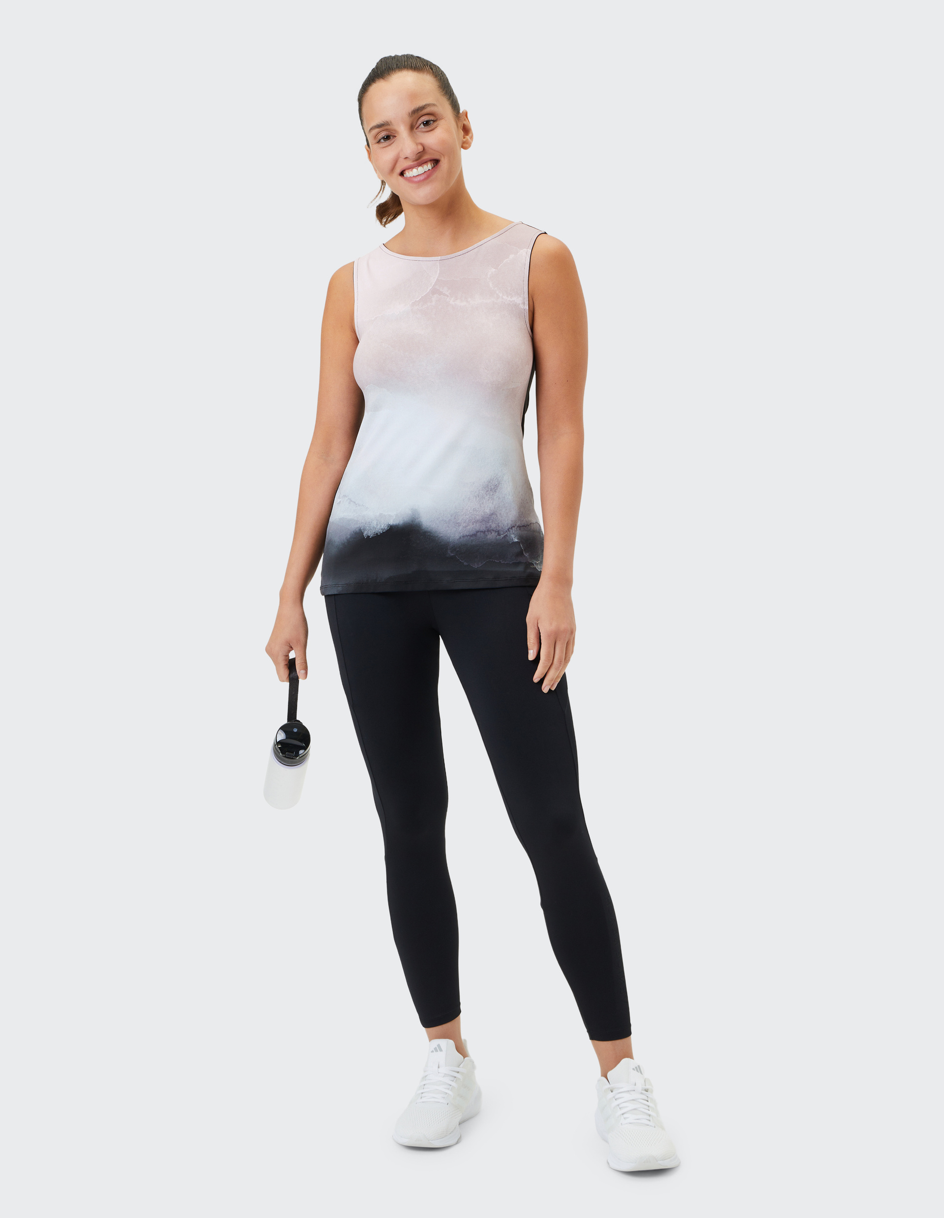 Frau in Sportkleidung lächelt, trägt ein ärmelloses Shirt, schwarze Leggings und hält eine Wasserflasche in der rechten Hand.