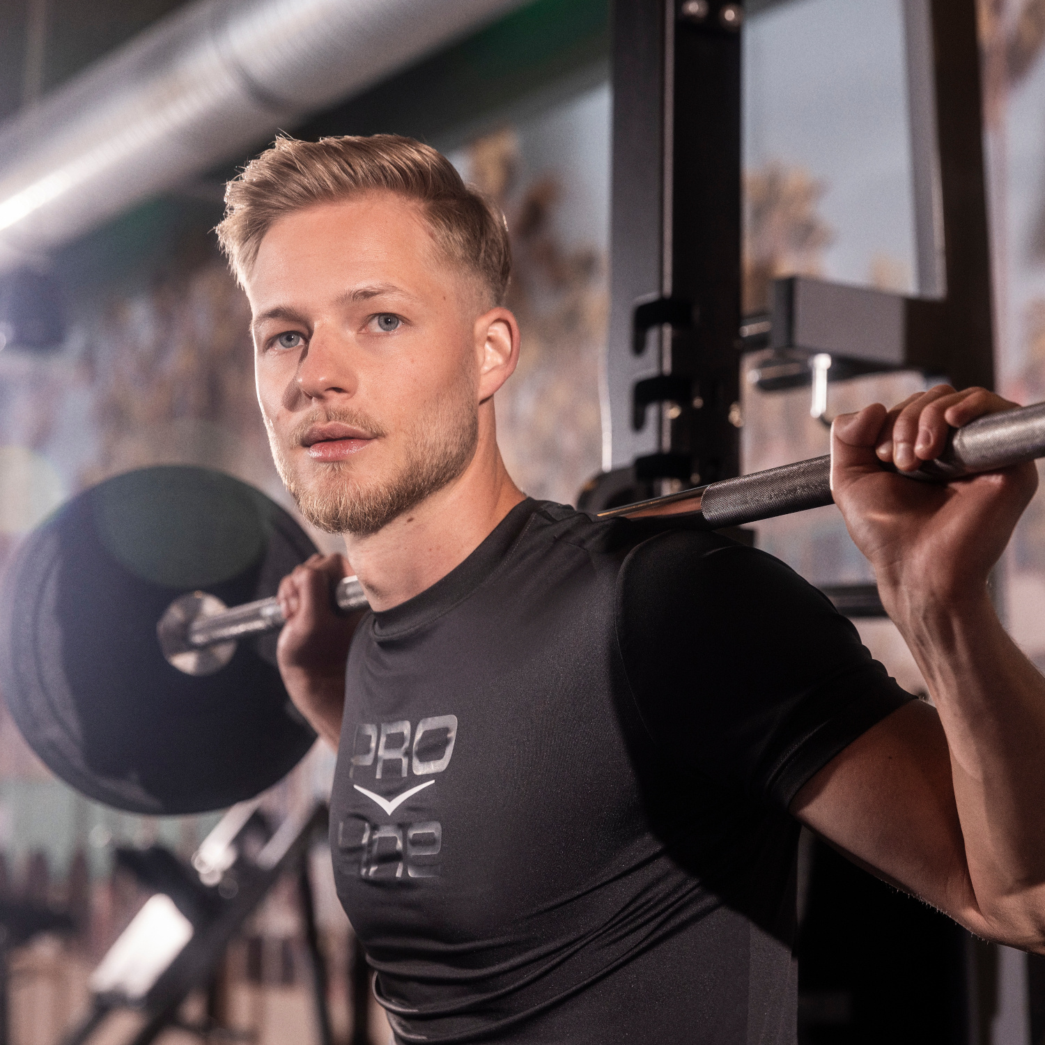 Ein Mann hebt im Fitnessstudio eine Langhantel auf den Schultern, trägt ein dunkles Sportshirt und blickt entschlossen in die Kamera.