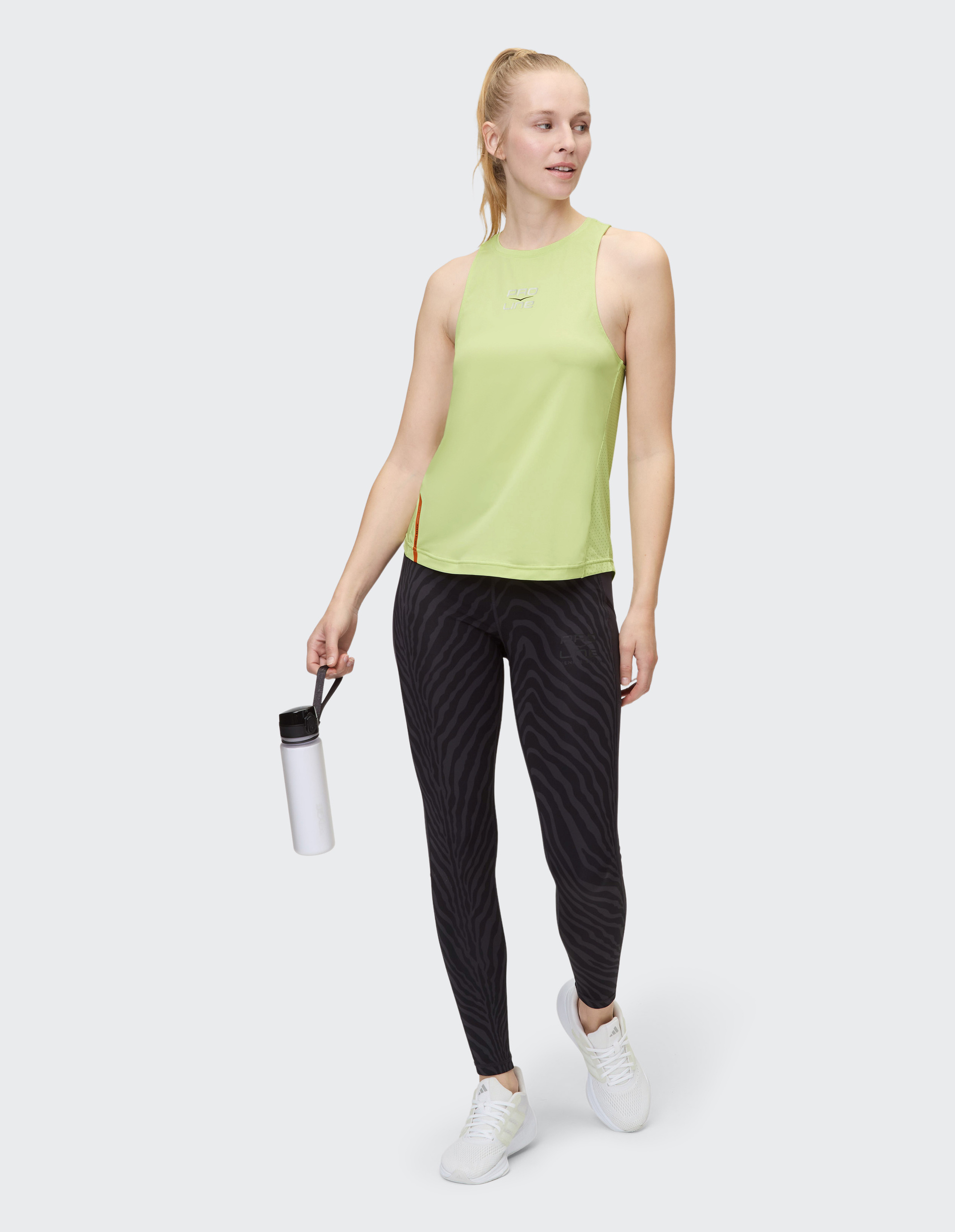Eine lächelnde Frau trägt ein grünes Tanktop, gestreifte Leggings und weiße Sneakers und hält eine weiße Trinkflasche.