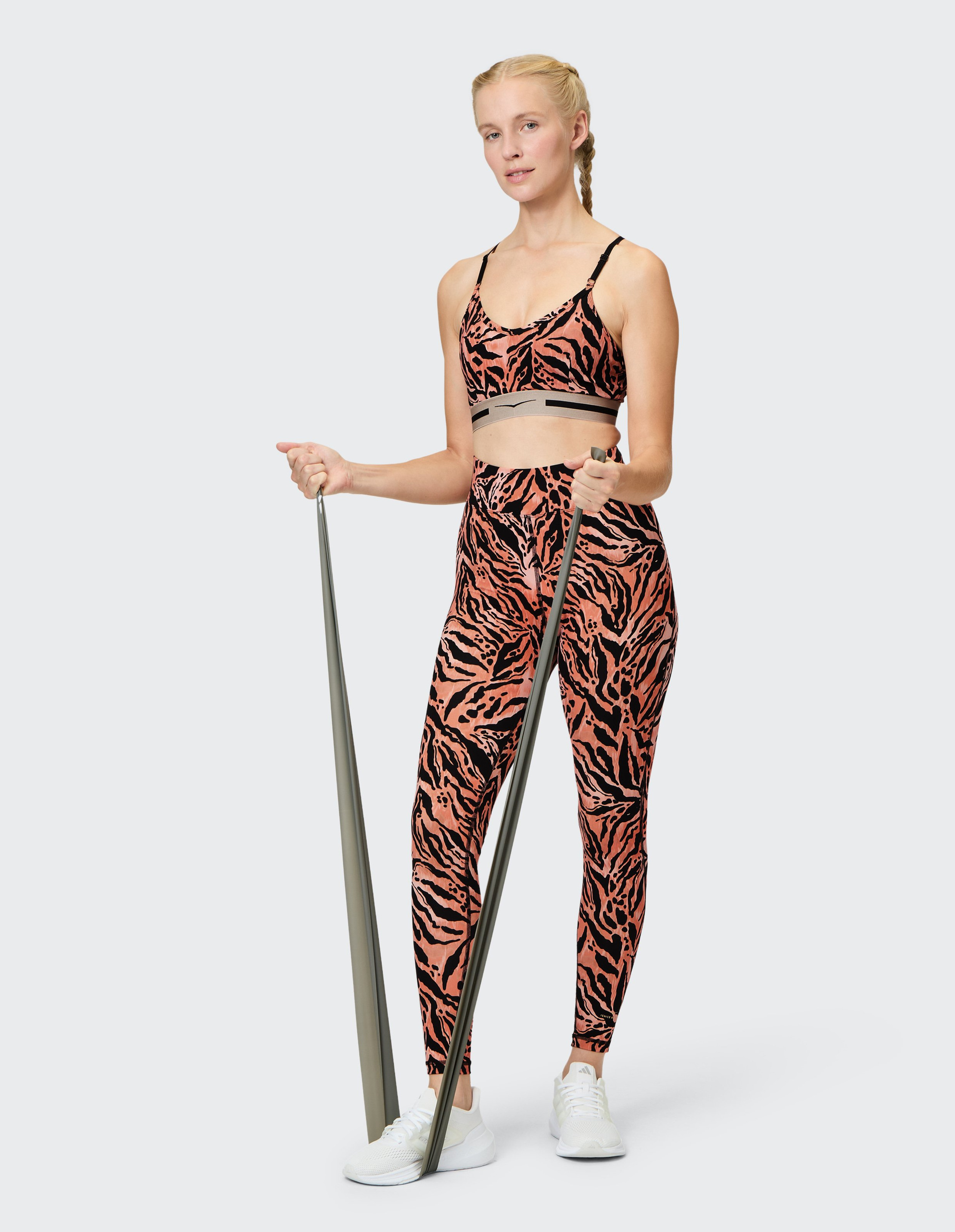 Frau in orange-schwarz gemustertem Sportoutfit steht mit Fitnessband in der Hand, lächelt leicht und trägt weiße Sportschuhe.