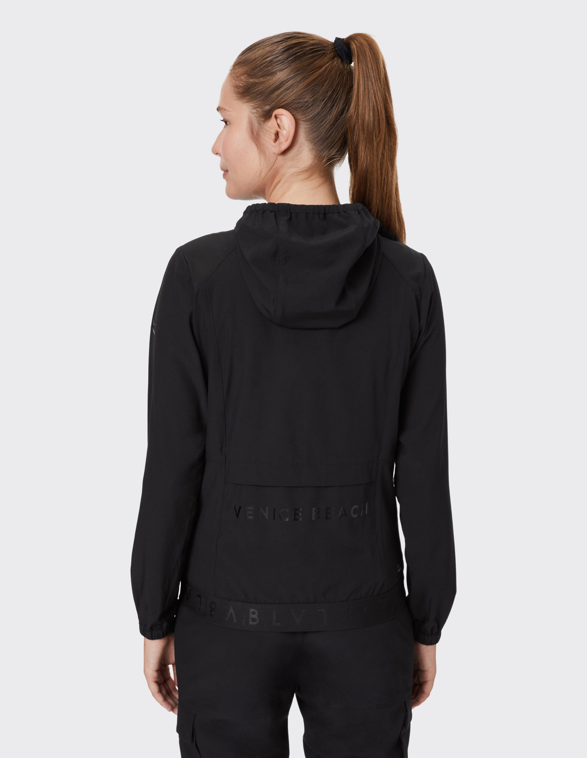 Eine Frau mit Pferdeschwanz trägt eine schwarze Kapuzenjacke mit dem Schriftzug "VENICE BEACH" auf der Rückseite.