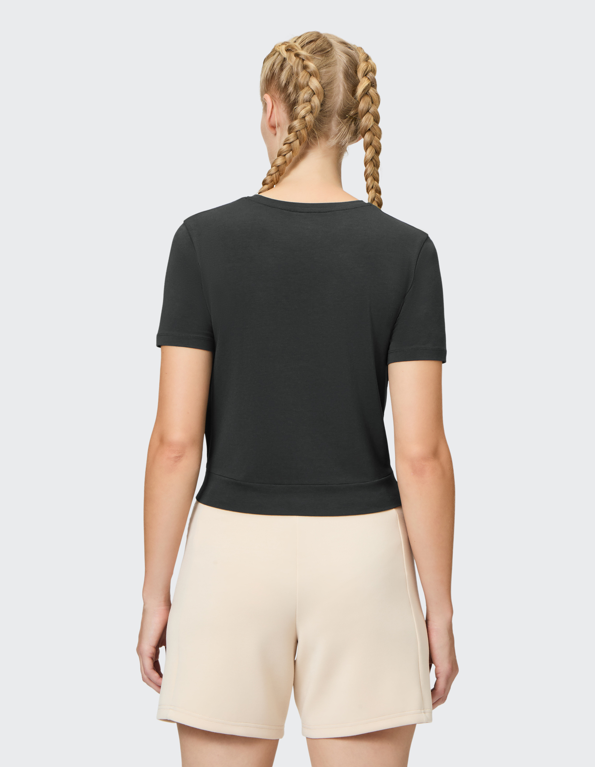 Rückansicht einer Person mit zwei geflochtenen Zöpfen, schwarzem T-Shirt und beigen Shorts vor grauem Hintergrund.