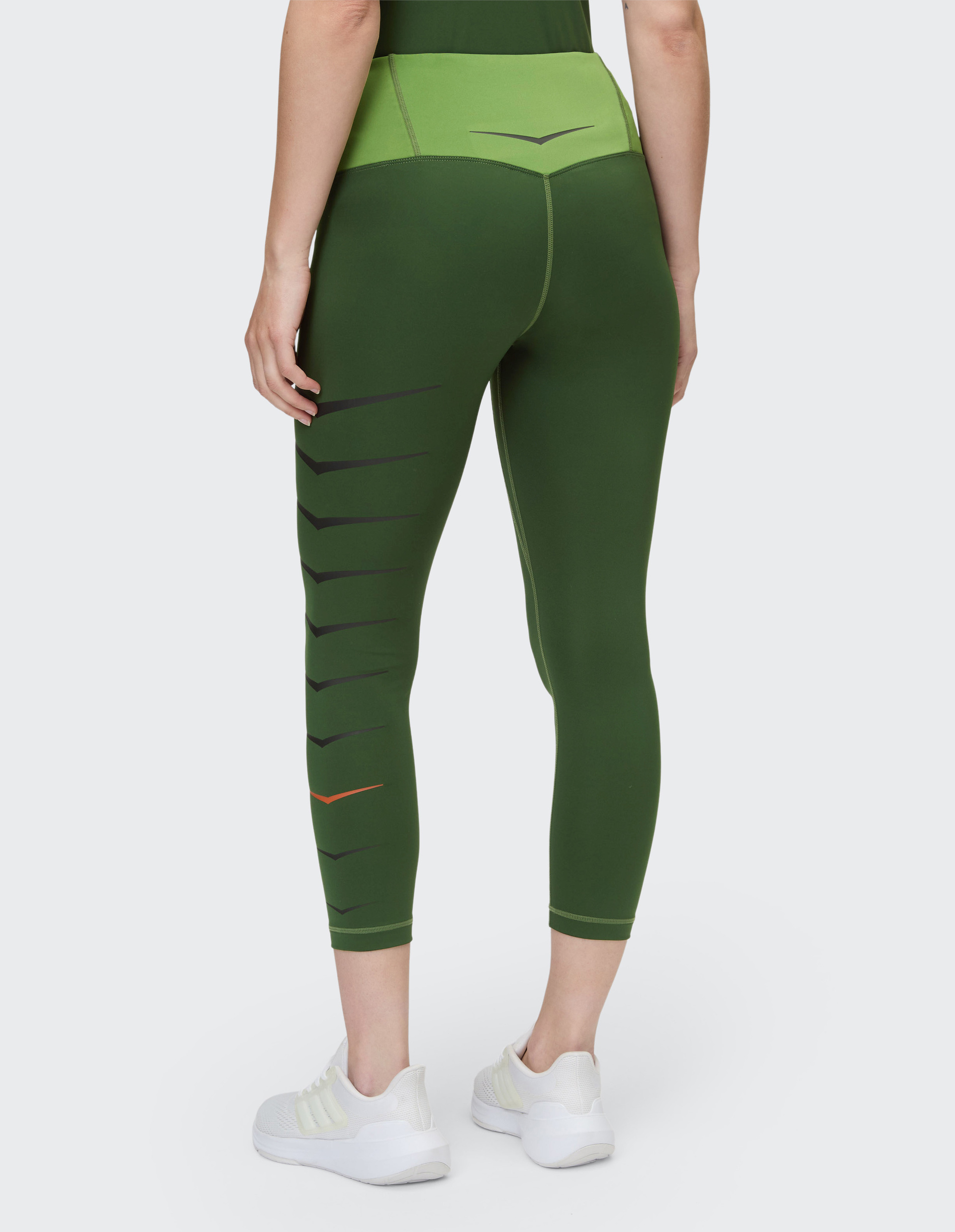 Person trägt grüne Leggings mit schwarzem Muster und leuchtender Y-Form am Bein, kombiniert mit weißen Turnschuhen auf grauem Hintergrund.