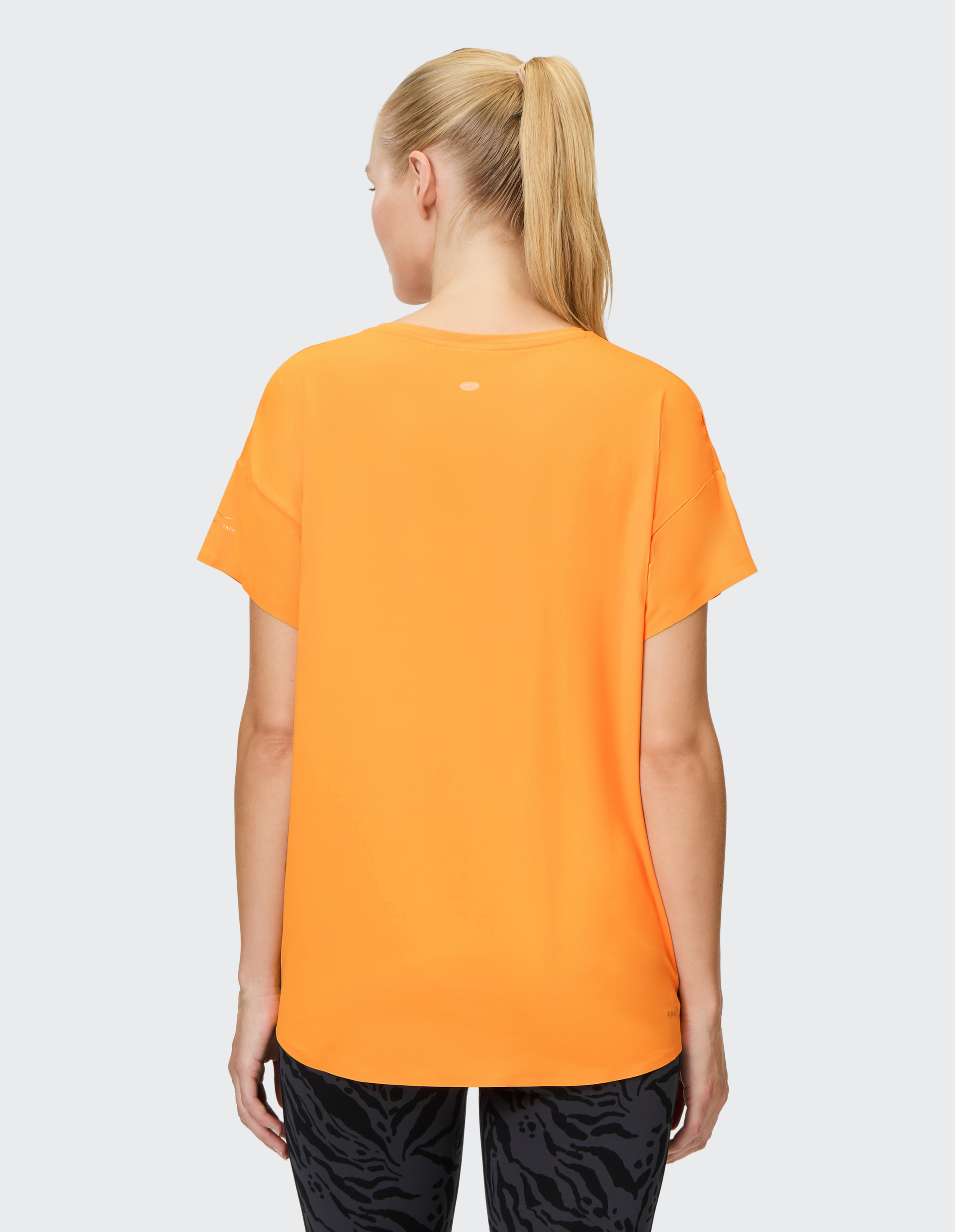 Frau mit Pferdeschwanz trägt ein leuchtend oranges T-Shirt und dunkle gemusterte Leggings, fotografiert von hinten vor grauem Hintergrund.