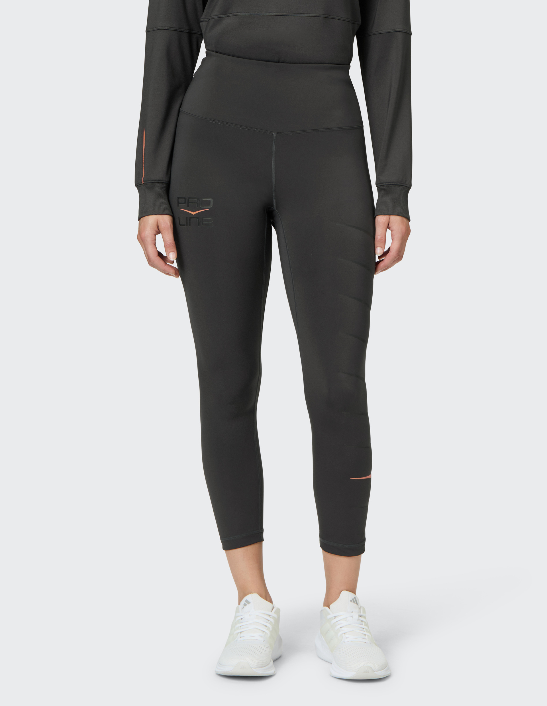 Frau trägt schwarze Leggings und Langarmshirt, mit weißen Sneakers, steht auf hellem Hintergrund, Logo "Pro Line" sichtbar.
