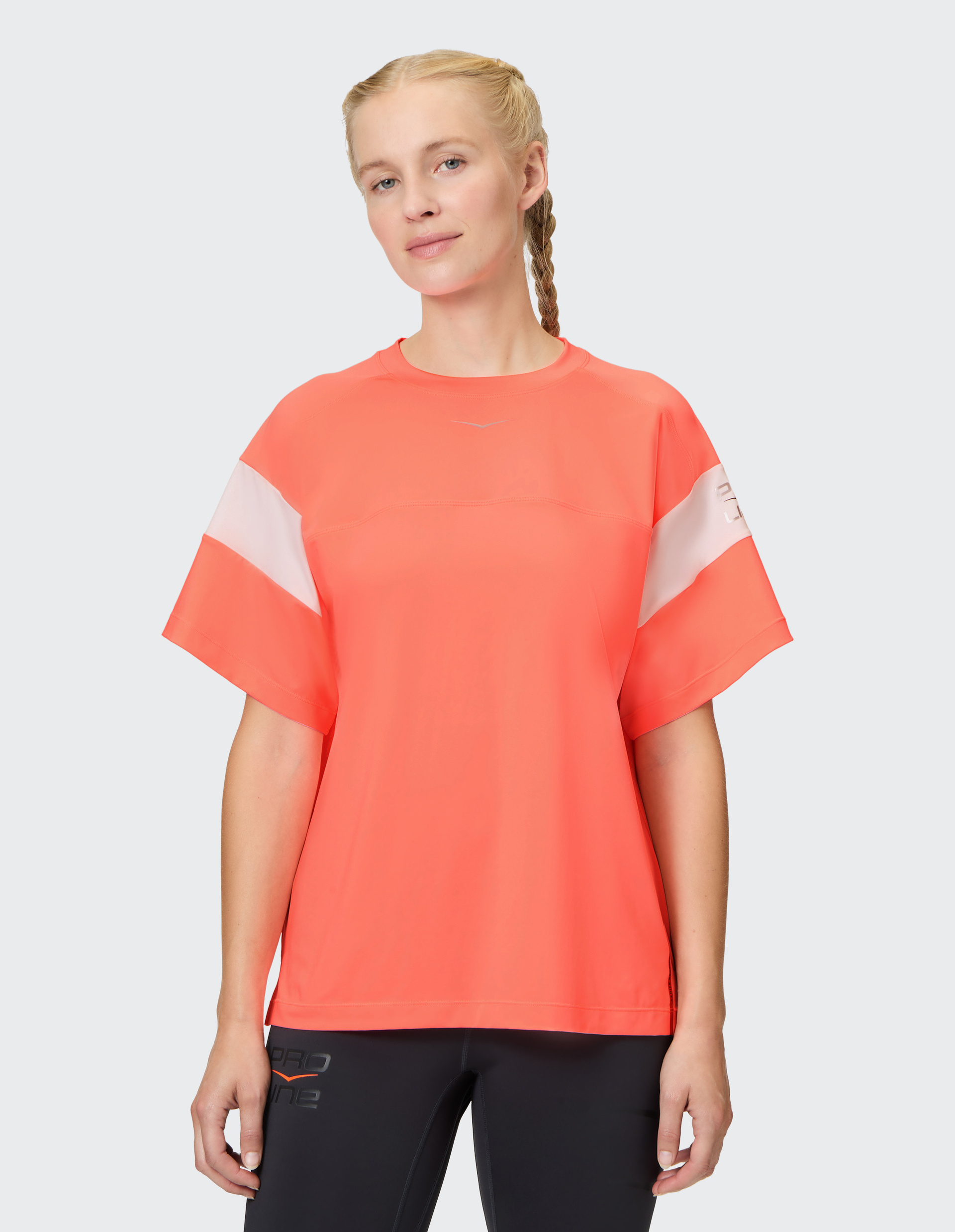 Person mit geflochtenem Haar trägt ein leuchtend oranges T-Shirt mit weißen Details und schwarze Hosen vor hellem Hintergrund.