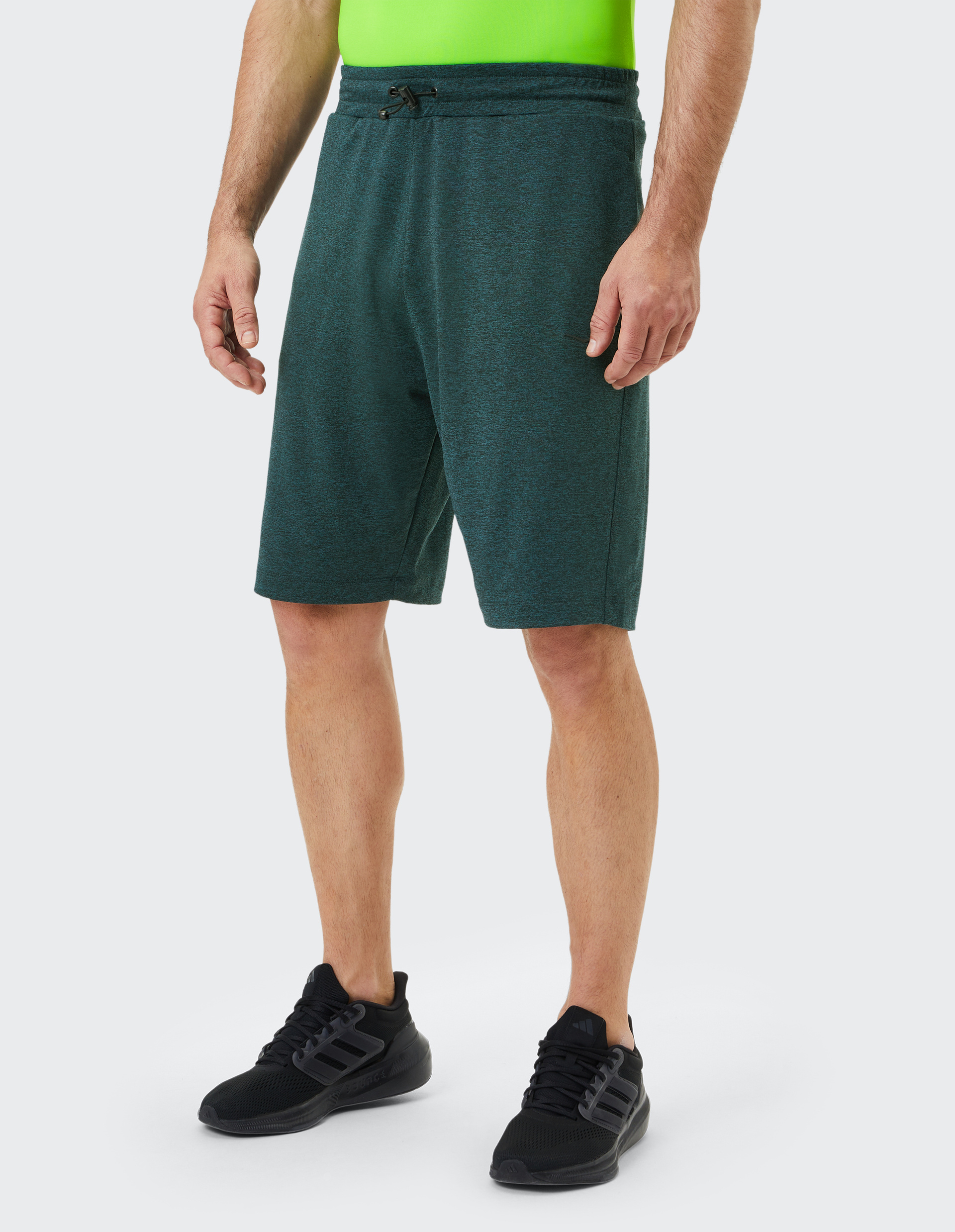 Mann trägt dunkle grüne Shorts mit Zugband und neonfarbenes Shirt, schwarze Sportschuhe auf hellem Hintergrund.